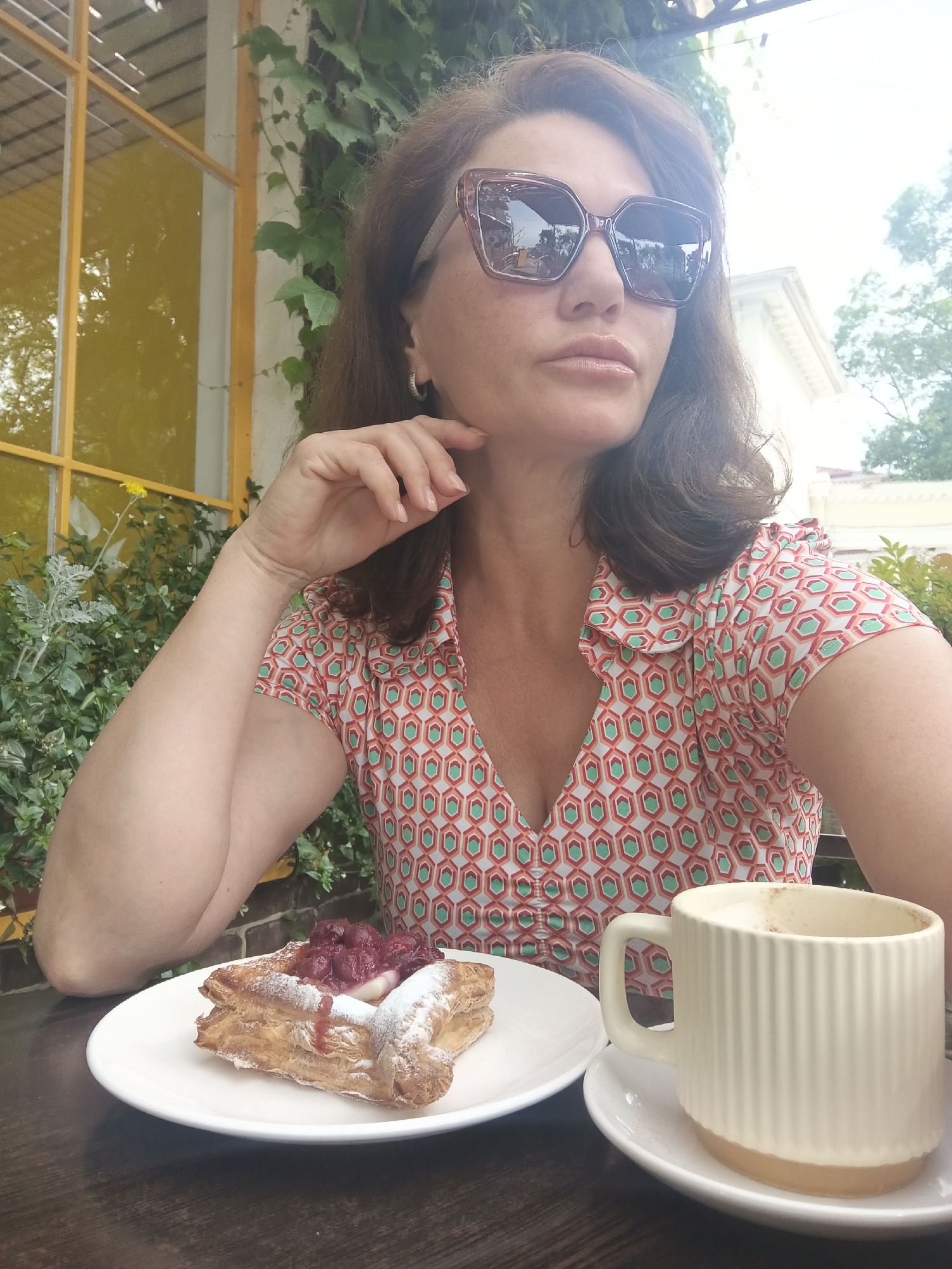 In Sochi, Russia, a woman with brown hair and large sunglasses is captured from the chest up, seated outdoors at a cafe. She wears a patterned coral and green blouse and rests her chin on her hand. In front of her on a white plate is a pastry topped with red fruit and cream, and to the right is a beige mug of coffee on a saucer. The background shows lush green foliage and a yellow-framed window, suggesting a vibrant, summery atmosphere.