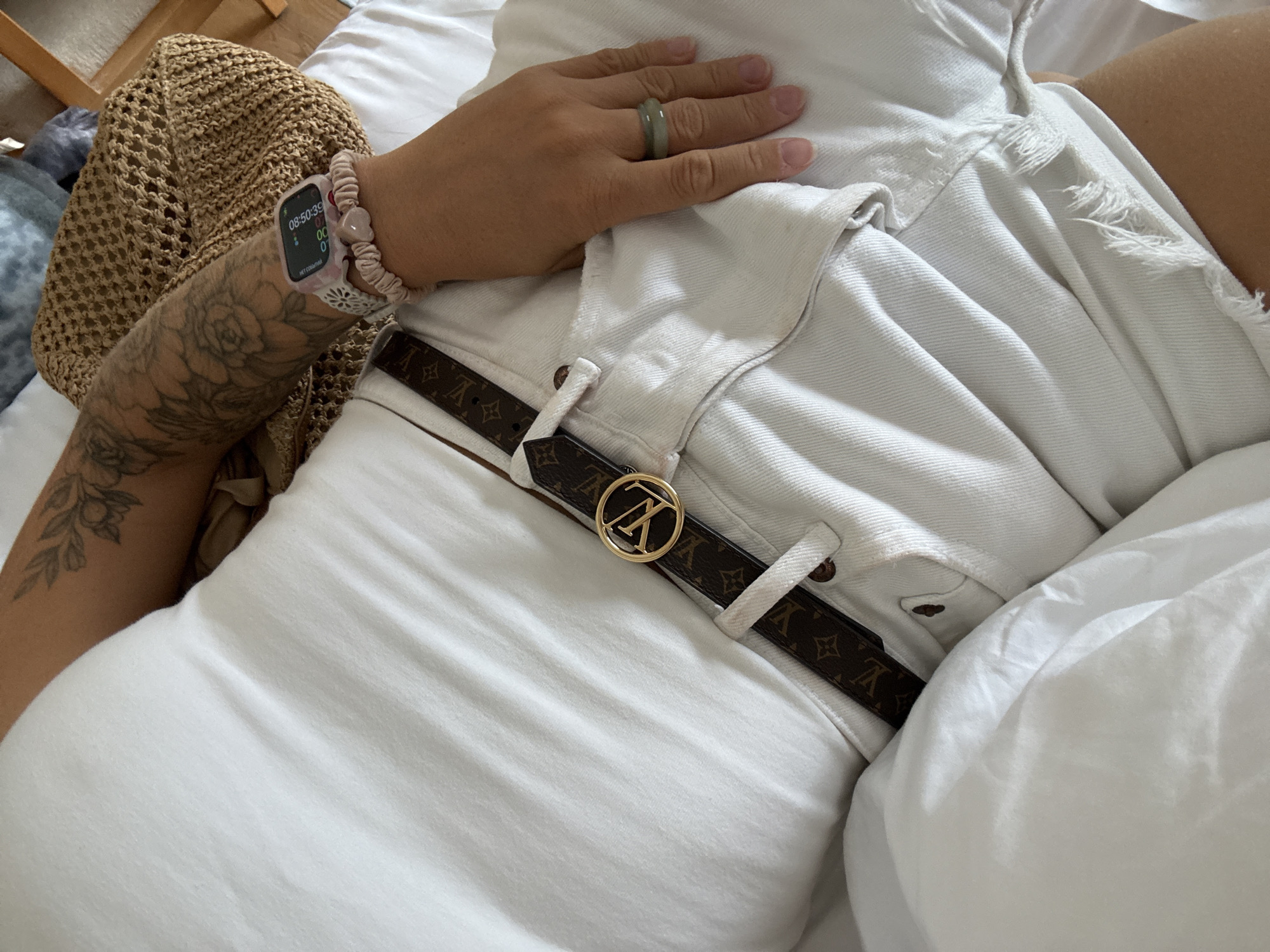 A person, lying on a bed covered by white sheets, is viewed from a high-angle, close-up perspective. The person is wearing white denim shorts, a brown Louis Vuitton belt with a gold buckle, and a white top. Their left arm, adorned with a floral tattoo, rests near a woven straw bag. On their wrist, a pink Apple Watch and a scrunchie with rose quartz beads are visible. Their right hand, wearing a green ring, is placed on their shorts. The scene is indoors, with soft lighting suggesting natural light. The specific location within Wuxing District, China, cannot be determined from the visual information provided.