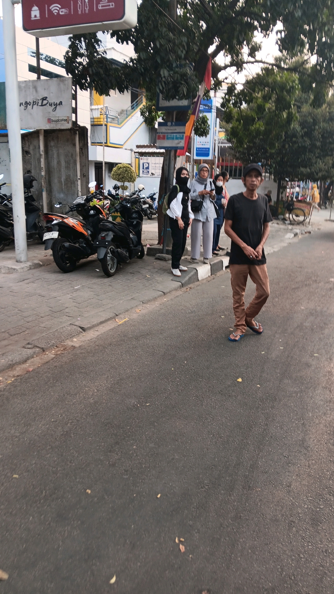 The image depicts a street scene in South Jakarta, Indonesia, likely in the vicinity of an area with commercial establishments, as indicated by signage. In the foreground, a man wearing a black t-shirt and brown pants with blue flip-flops stands facing the camera on an asphalt road. Behind him, a sidewalk features several parked motorcycles, predominantly black and orange. Three individuals are standing on the sidewalk, two women wearing hijabs, one in a black and white outfit and the other in a light grey outfit, and a third person in a blue shirt. One woman is looking towards the camera, while the other two are looking away. A building with yellow and white accents is visible in the background, along with trees and utility poles. Further down the street, a vendor cart is visible. The lighting suggests it is either early morning or late afternoon.
