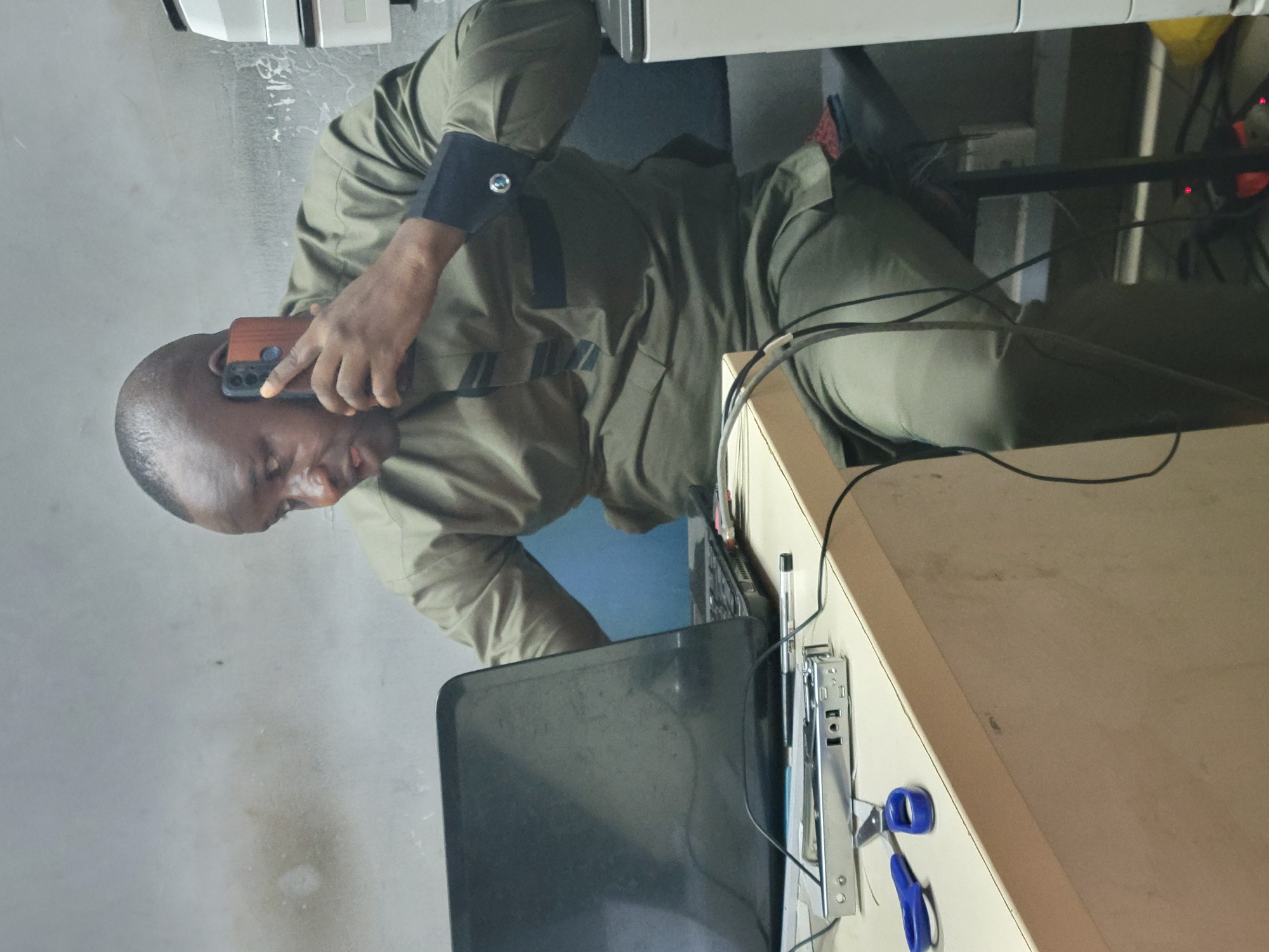 The image shows a man in Abuja, Nigeria, sitting at a desk and holding a mobile phone to his ear. He wears a light brown long-sleeved shirt with dark cuffs. A laptop sits open on the desk in front of him, along with a stapler and a pair of scissors. There is a computer tower case behind the laptop. The background consists of a wall and some office equipment. Wires are visible around the desk and computer.