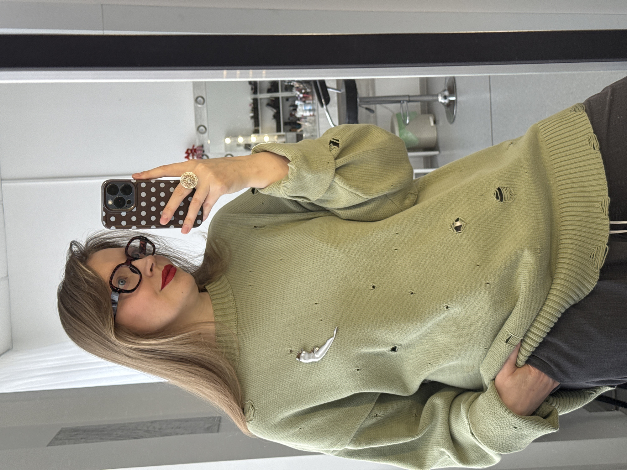 A woman is taking a selfie in front of a mirror. She has long blonde hair and wears glasses and red lipstick. She is wearing a light green, distressed sweater with holes and a silver brooch. She is holding a smartphone with a brown and white polka dot case. She is wearing a large gold ring on her right ring finger. In the background, a makeup vanity with lights and various items on shelves can be seen. The location of the image is unknown, and it is not possible to determine if it is in Izhevsk, Russia.