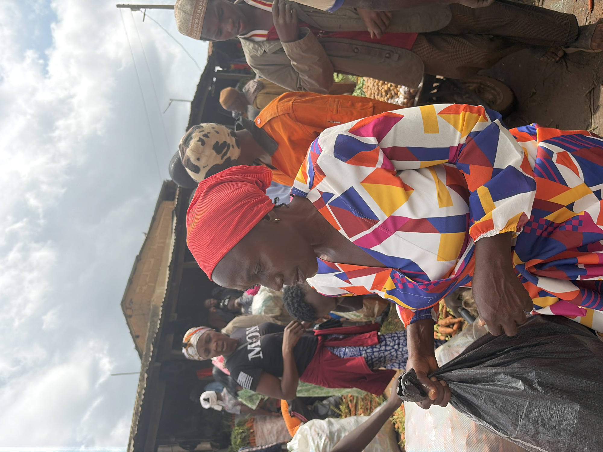 A medium shot captures a busy outdoor market scene in Gura Top, Nigeria, under a partly cloudy sky. In the foreground, a woman wearing a vibrant, geometric-patterned dress and a red headscarf is holding a black plastic bag. Her hands are visible as she grasps the bag. To her left, another person's arm is visible, holding a black object. In the mid-ground, several individuals are present. One woman wears a black t-shirt with the word "MON" printed on it and a red garment tied around her waist. Another person is seen in an orange jumpsuit. Behind them, a man in a patterned cap and another in a tan jacket and cap are visible. Stalls with awnings are set up in the background, with piles of produce, possibly carrots, visible on the ground. The ground of the market appears to be dirt.