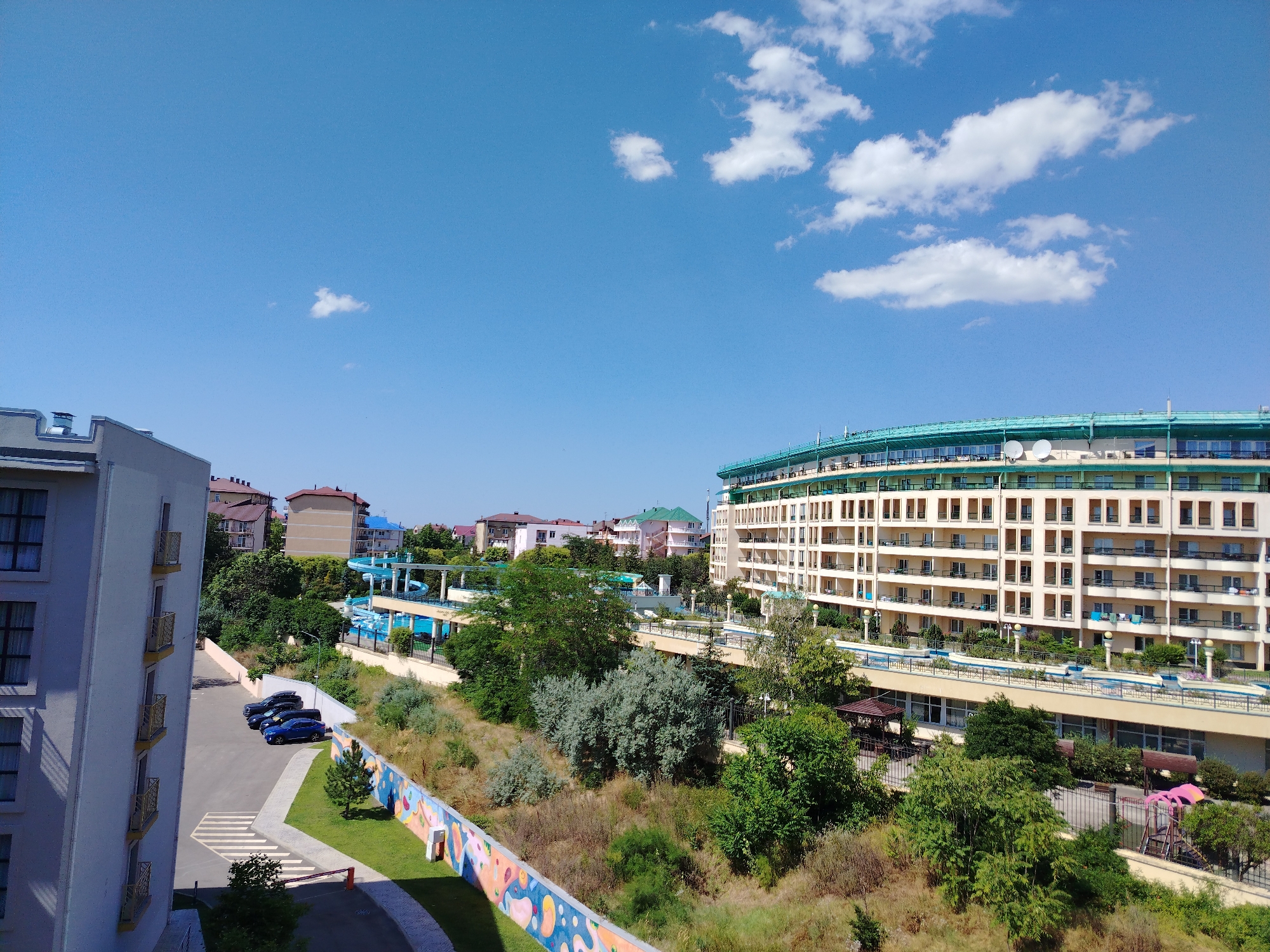 The image captures a bright, sunny day in an urban area, likely a resort or hotel complex, with a clear blue sky dotted with scattered white clouds. In the foreground on the left, a tall, light-colored building with multiple balconies and windows is visible, suggesting residential or hotel accommodations. Adjacent to it is a parking lot with three cars, two dark-colored SUVs and a blue sedan. A pedestrian crosswalk is marked on the asphalt.
Dominating the right side of the image is a large, multi-story building with a curved facade, featuring numerous balconies and windows, and a distinctive teal-colored roofline. This building appears to be a hotel or apartment complex. Below this main building, a blue-tiled swimming pool and water slides are visible, indicating a recreational area.
The middle ground is characterized by a mix of lush green trees, shrubs, and dry, overgrown vegetation, suggesting a natural or landscaped area separating different parts of the complex. A colorful mural adorns a low wall in this section, adding a vibrant visual element.
In the background, several other buildings with pitched roofs, some in shades of pink and green, can be seen, contributing to the urban landscape. The overall impression is one of a well-maintained resort or residential area on a clear summer day.