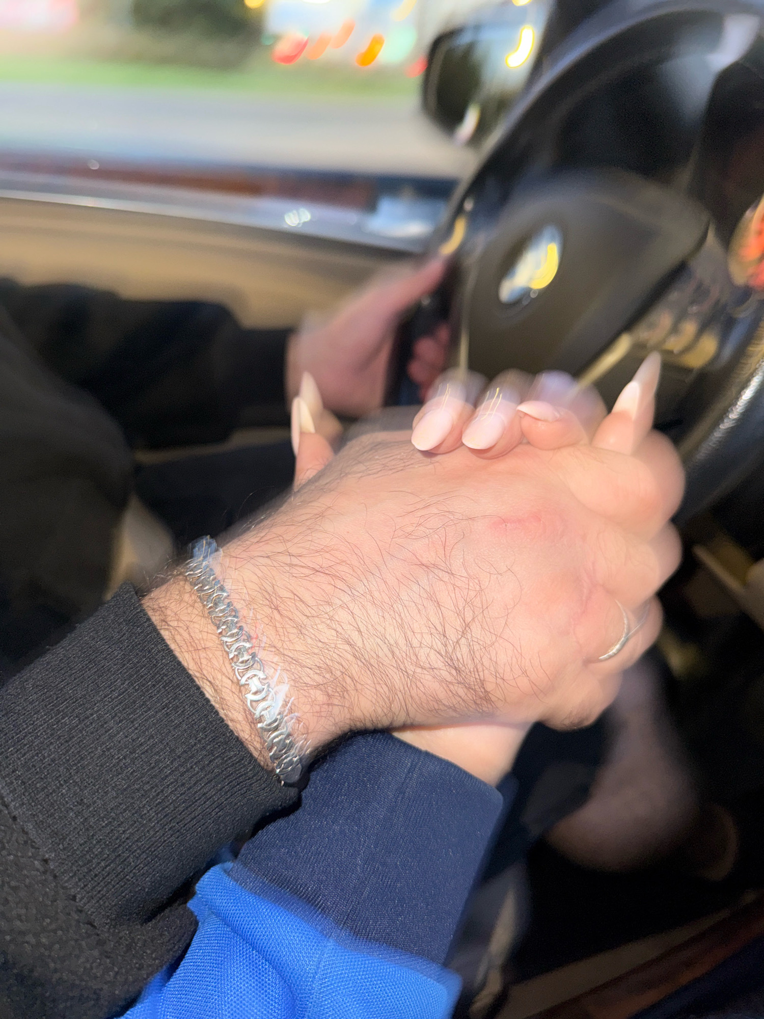 Two individuals are holding hands while seated in a car. One individual, wearing a black long-sleeved shirt, has a silver chain bracelet on their wrist. The other individual, with long, light-colored fingernails, wears a ring on their left hand. The background shows blurred lights, suggesting the car is moving or parked at night in an urban environment. There are no specific visual cues to determine the location within Tallinn, Estonia.