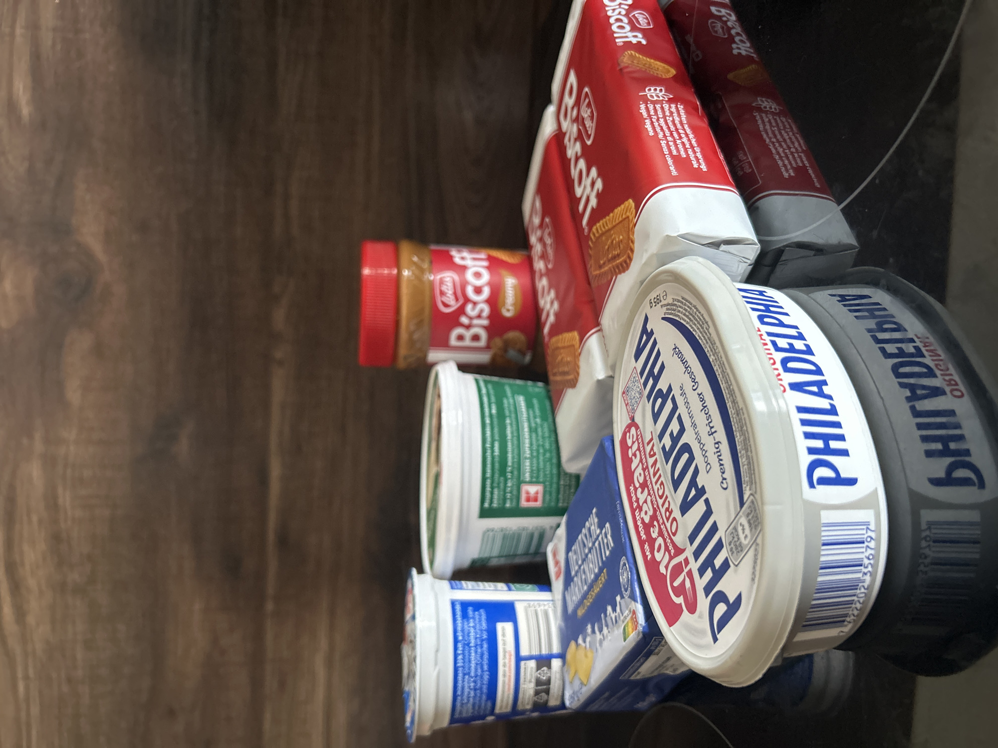 A variety of food products are arranged on a dark, reflective surface, likely a stovetop, against a dark wood-grain background. In the foreground, a tub of Philadelphia Original cream cheese is prominently displayed, with its blue and white labeling clearly visible. To the left of the cream cheese is a blue package of what appears to be German butter, labeled "DEUTSCHE MARKDBITTER". Behind these items, to the left, is a white plastic tub with blue labeling, possibly yogurt. To its right is another white tub with green labeling, also likely a dairy product. In the middle ground, to the right, are two red packages of Lotus Biscoff cookies. Behind the cookies and the tubs is a small jar of Lotus Biscoff spread with a red lid. The lighting is direct, creating reflections on the surface. No people are present in the image.
