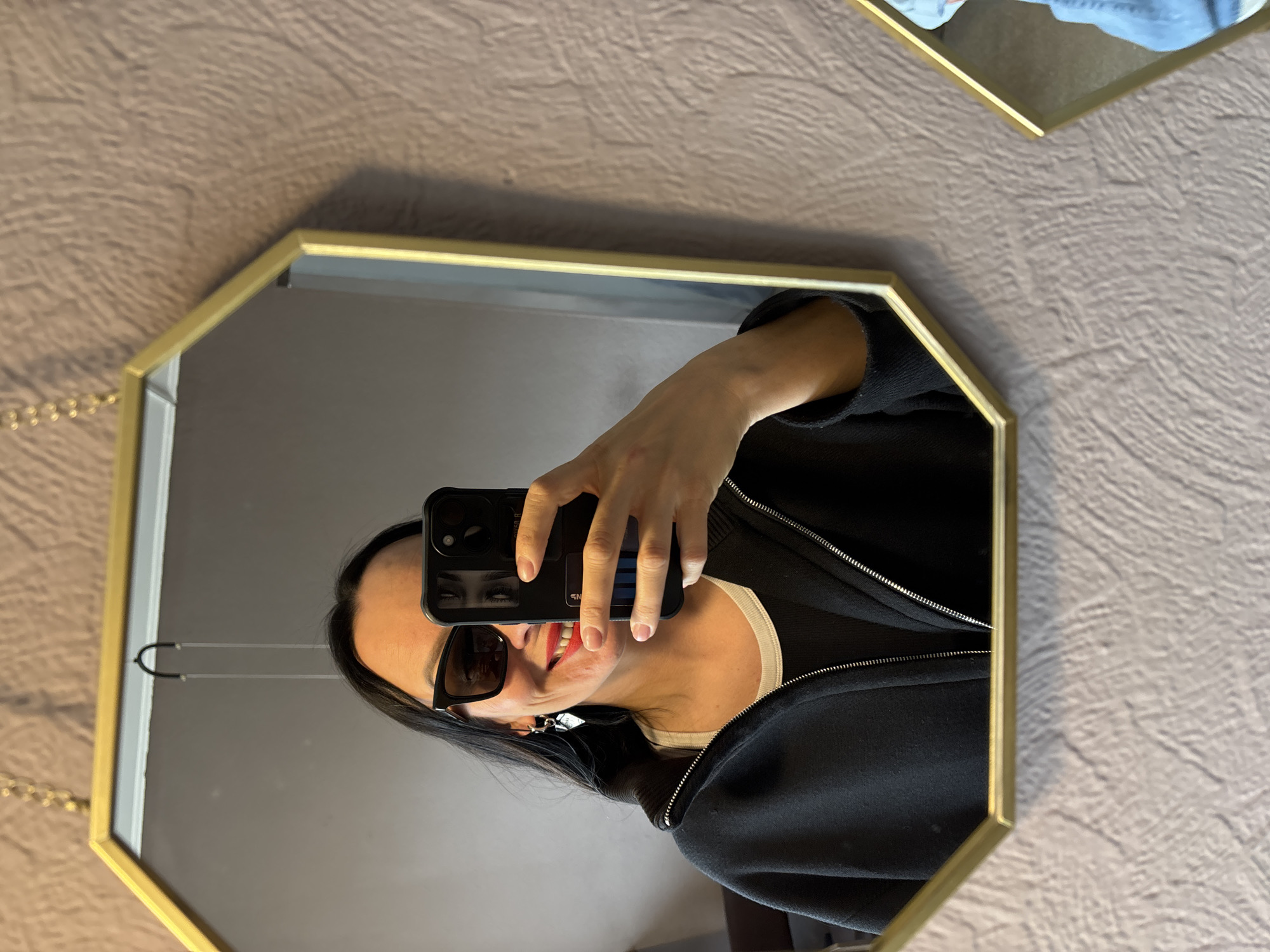 A person is taking a selfie in an octagonal mirror with a gold frame. The person is wearing sunglasses, a black zip-up jacket, and a white top underneath. The phone is held up to capture the reflection. The background is a light brown textured wall. The location details (city of Chaikovsky, Russia) are not discernible from the image.