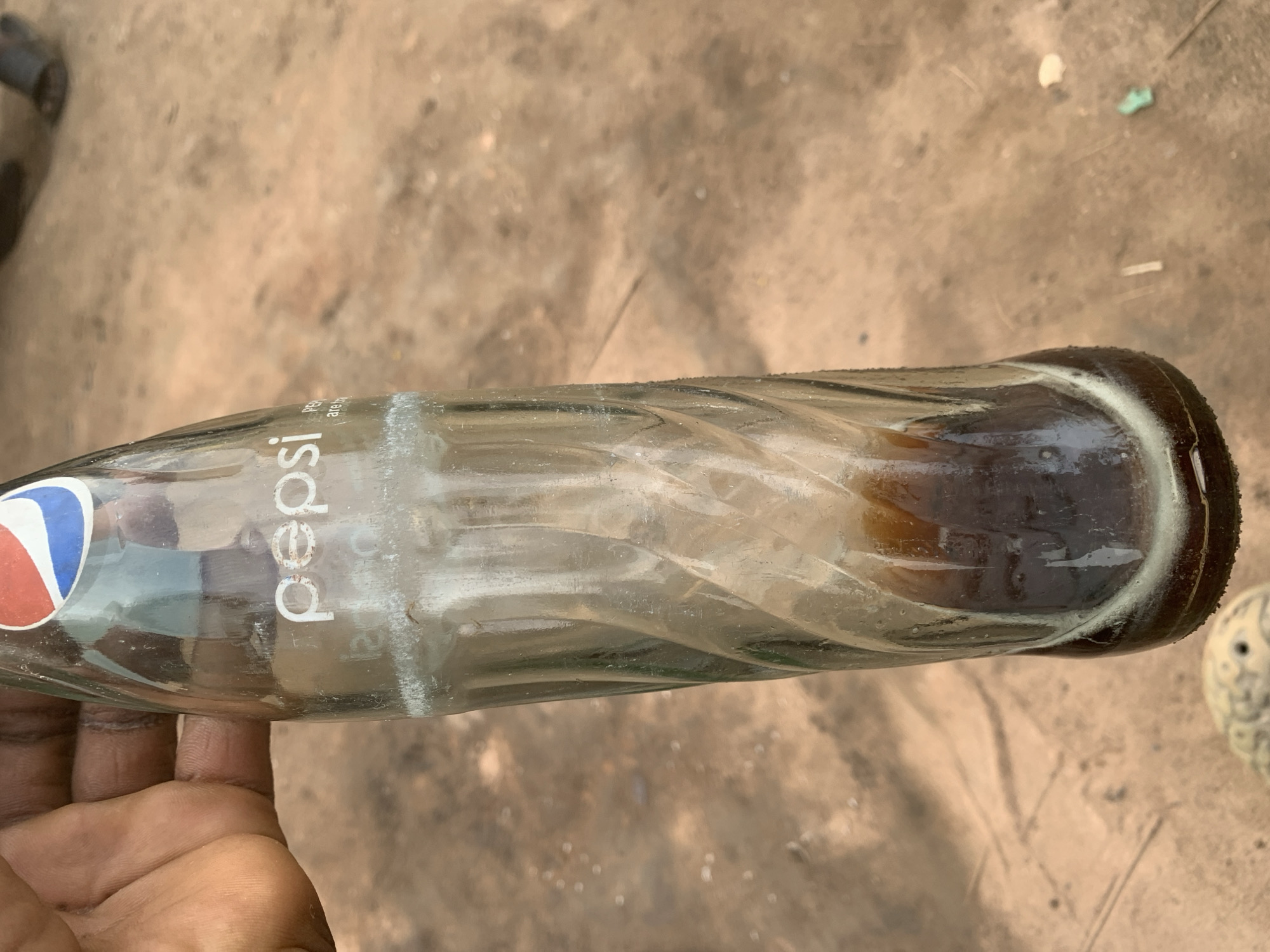 A close-up, eye-level shot shows a hand holding a clear glass bottle of Pepsi, which contains a dark brown liquid. The bottle is partially filled with the drink, and there are some white residues on the inside. The bottle has a ribbed design. The background is a dry, sandy ground, suggestive of an outdoor environment. In the blurred background, a person's legs and feet, wearing dark sandals, are visible. The overall impression is one of a casual, perhaps informal setting.
