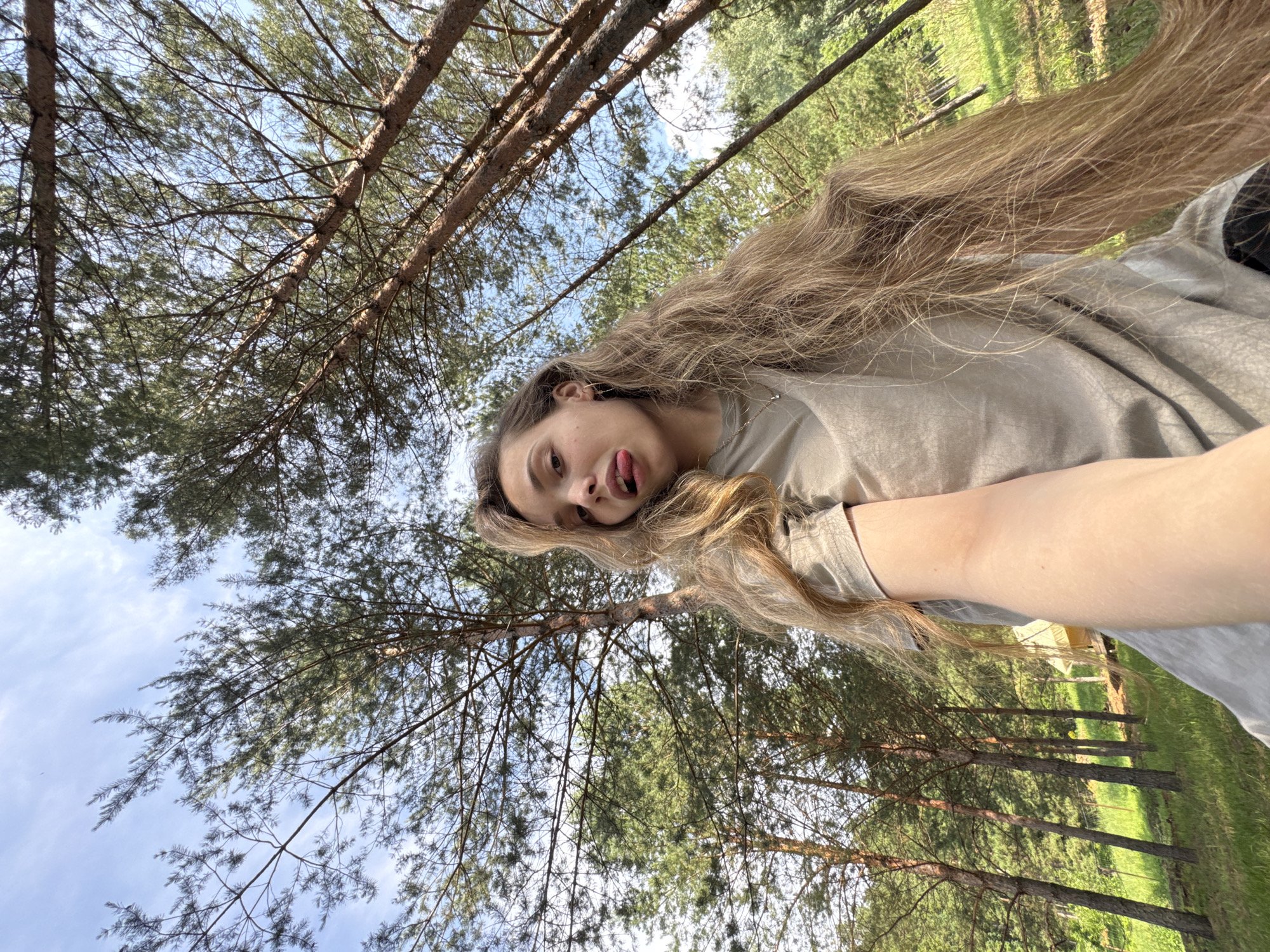 A young woman with long, blonde, wavy hair is taking a selfie outdoors in a forest. She is wearing a plain, light-colored t-shirt and has her tongue sticking out playfully. The background consists of tall, slender pine trees against a bright blue sky with some wispy clouds. The ground is covered in green grass, and a yellow tent-like structure is visible in the distance, partially obscured by trees. The setting appears to be a campground or a wooded recreational area. There is no specific indication of the city of Гольяны, Russia, in the image itself.