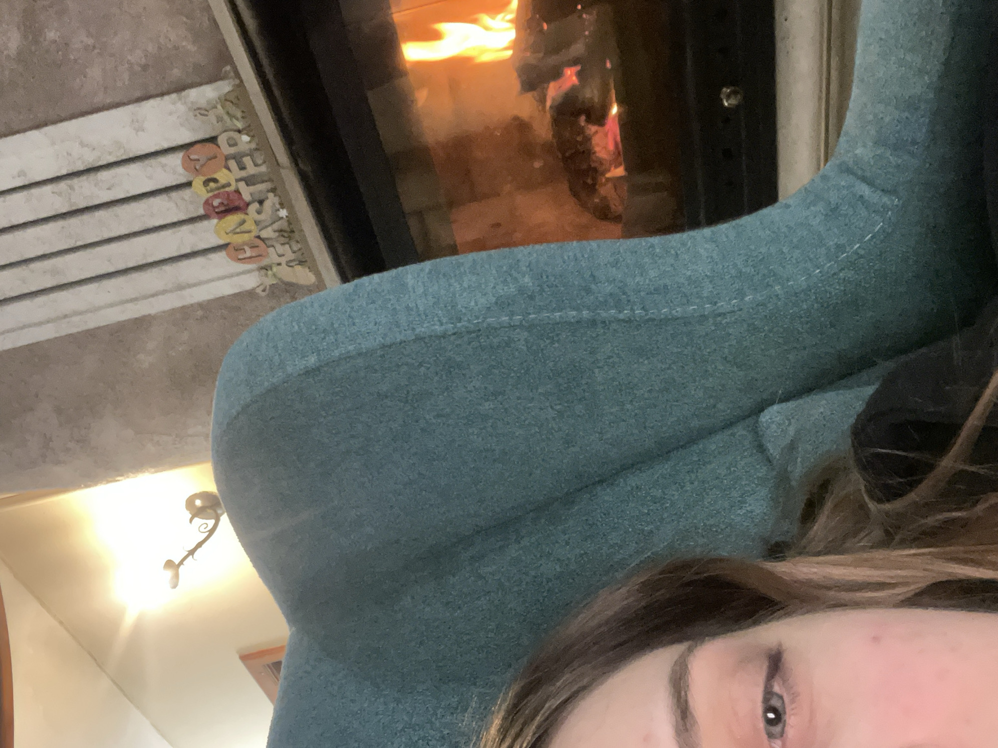 A close-up, tilted indoor shot focuses on a person's face, specifically their eye and forehead, with brown and blonde hair. The person is positioned in front of a teal-colored upholstered object, possibly a chair. Behind the chair is a fireplace with a visible fire. To the left of the fireplace is a white surface with the word "EASTER" spelled out using decorative letters/eggs. Above the person is a wall-mounted light fixture with a curved design. The location is not discernible from the image and cannot be specified as Tsigov chark, Bulgaria.