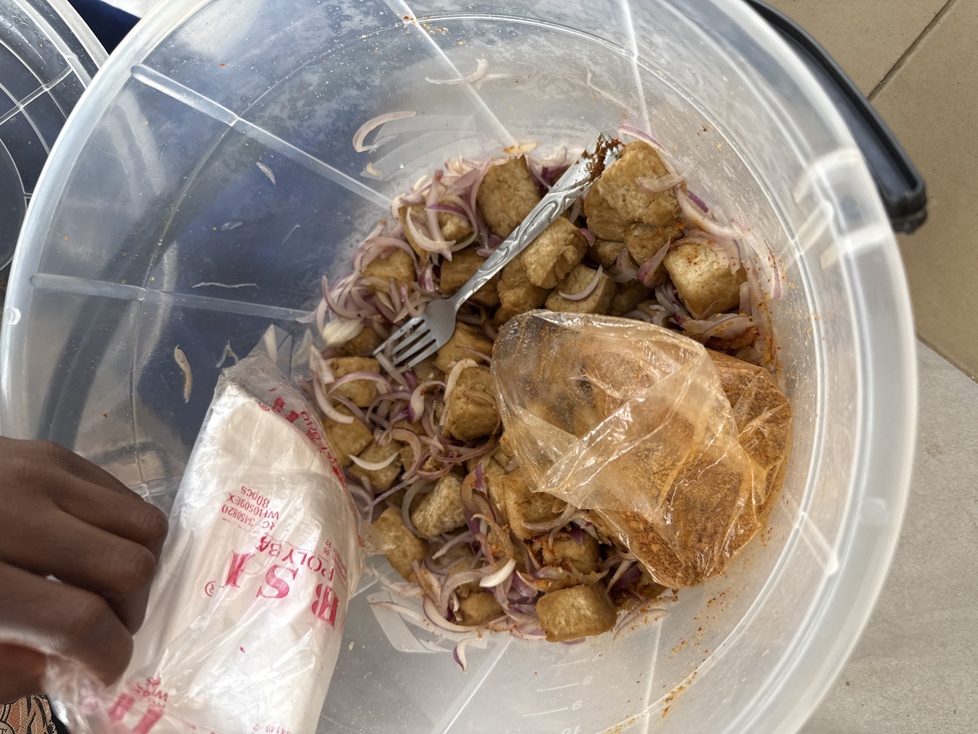 In Abuja, Nigeria, a close-up overhead shot captures a large, clear plastic basin filled with food. The basin contains numerous cubed pieces of fried tofu, thinly sliced red onions, and a small plastic bag filled with a reddish-brown powder, likely a spice mix. A silver fork rests on top of the food. A hand, partially visible at the top left, is holding a white plastic bag with red lettering that reads "POLY BAG." The lighting suggests an outdoor or well-lit indoor environment.