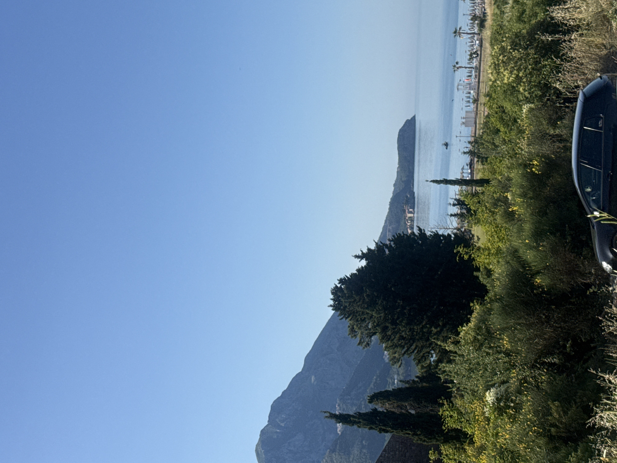 This wide-angle outdoor photograph captures a sunny coastal landscape in Boreti, Montenegro. The upper half of the image is dominated by a clear, unblemished blue sky, indicating fair weather.
In the foreground, the lower portion of the image is occupied by dense, varied vegetation. This includes numerous green shrubs, some displaying small yellow flowers, interspersed with areas of dried, light brown grasses. On the far left, a partial stone structure is visible. Towards the center-left, a cluster of tall, slender conifer trees, resembling cypresses, stands prominent, alongside a larger, dark green, bushy conifer tree slightly to their right. The rear-side profile of a dark-colored car, consistent with a hatchback or wagon, is partially visible at the bottom right.
The mid-ground features a calm, light blue-green sea extending from the left to the right. A small, dark-colored boat is positioned in the water near the center. To the right, a developed beach area is visible, equipped with multiple rows of white beach umbrellas and accompanying sun loungers. Several tall palm trees are scattered along the beach or adjacent grassy sections, and a white, multi-level structure resembling a lifeguard tower is situated on the sand. A prominent headland or promontory extends into the sea from the far right side of the frame, with distant buildings discernible along the coast at its base.
The background on the left is dominated by a large, rugged mountain range, appearing dark blue-grey. The mountains exhibit discernible slopes and some lighter patches indicative of exposed rock or sparse vegetation. Small white settlements or structures are visible on the lower mountain slopes. No individuals are clearly discernible within the scene, and the car and boat appear stationary.