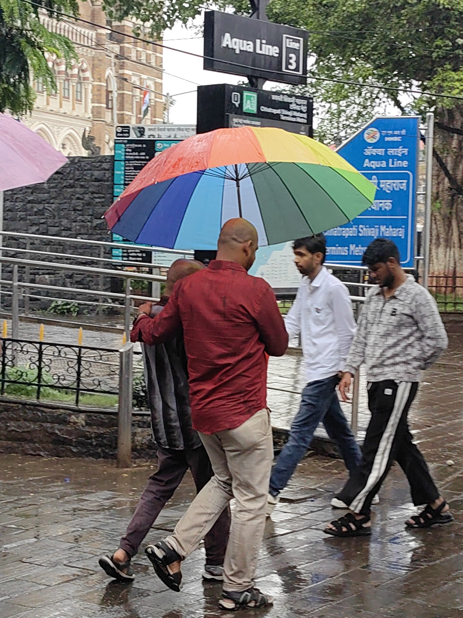 A group of four men are walking on a wet, paved sidewalk in Mumbai, India, under a colorful, open umbrella. The man in the foreground, facing away from the camera, wears a red long-sleeved shirt and light-colored pants. He is holding the umbrella over himself and another man who is walking beside him, partly obscured. This second man is wearing a dark, possibly gray, long-sleeved shirt and dark pants. To their right, a third man in a white long-sleeved shirt and blue jeans walks ahead, while a fourth man in a patterned light-colored shirt and black pants with a white stripe walks slightly behind. The ground is wet and reflective, suggesting it is raining. In the background, a large building with intricate architecture, possibly a heritage building like the Chhatrapati Shivaji Maharaj Terminus, is visible, along with signage in Hindi and English indicating "Aqua Line" and "Line 3". Trees with lush green foliage are also present, contributing to the urban park-like atmosphere. The scene appears to be taking place during daytime, with overcast lighting conditions.