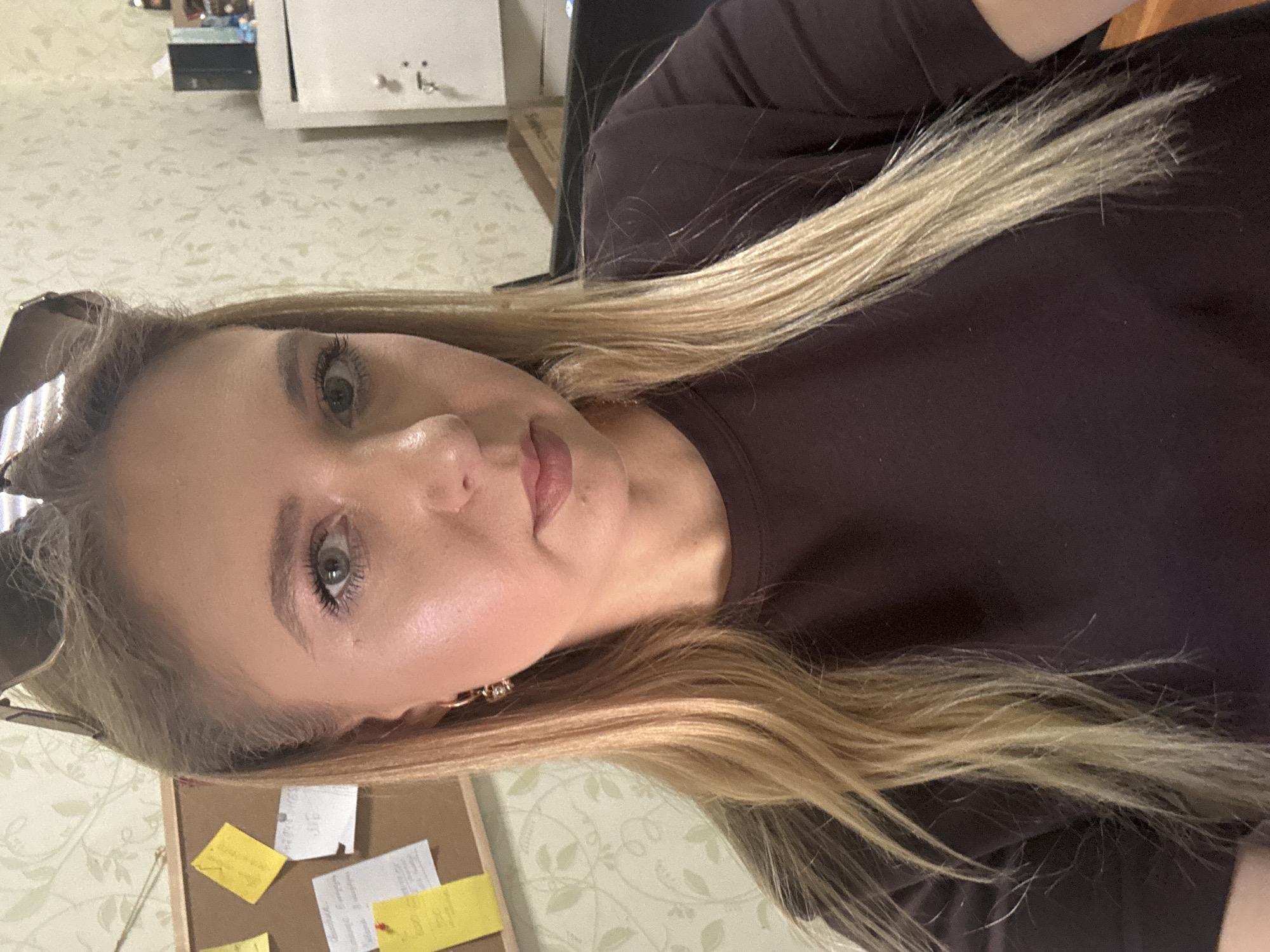 A close-up, head-and-shoulders selfie of a young, Caucasian woman with long, blonde hair and light-colored eyes. She is wearing a dark brown, crew-neck t-shirt. Her hair is styled down, with some strands falling over her shoulders. She has on light makeup, including mascara, eyeliner, and lipstick. She is also wearing small, hoop earrings. Her sunglasses are resting on top of her head. The background appears to be an indoor setting, possibly an office or study, with a corkboard on the left displaying several yellow and white sticky notes. To the right, there is a patterned wallpaper and a white cabinet with a lock. The lighting is bright, highlighting the woman's face.