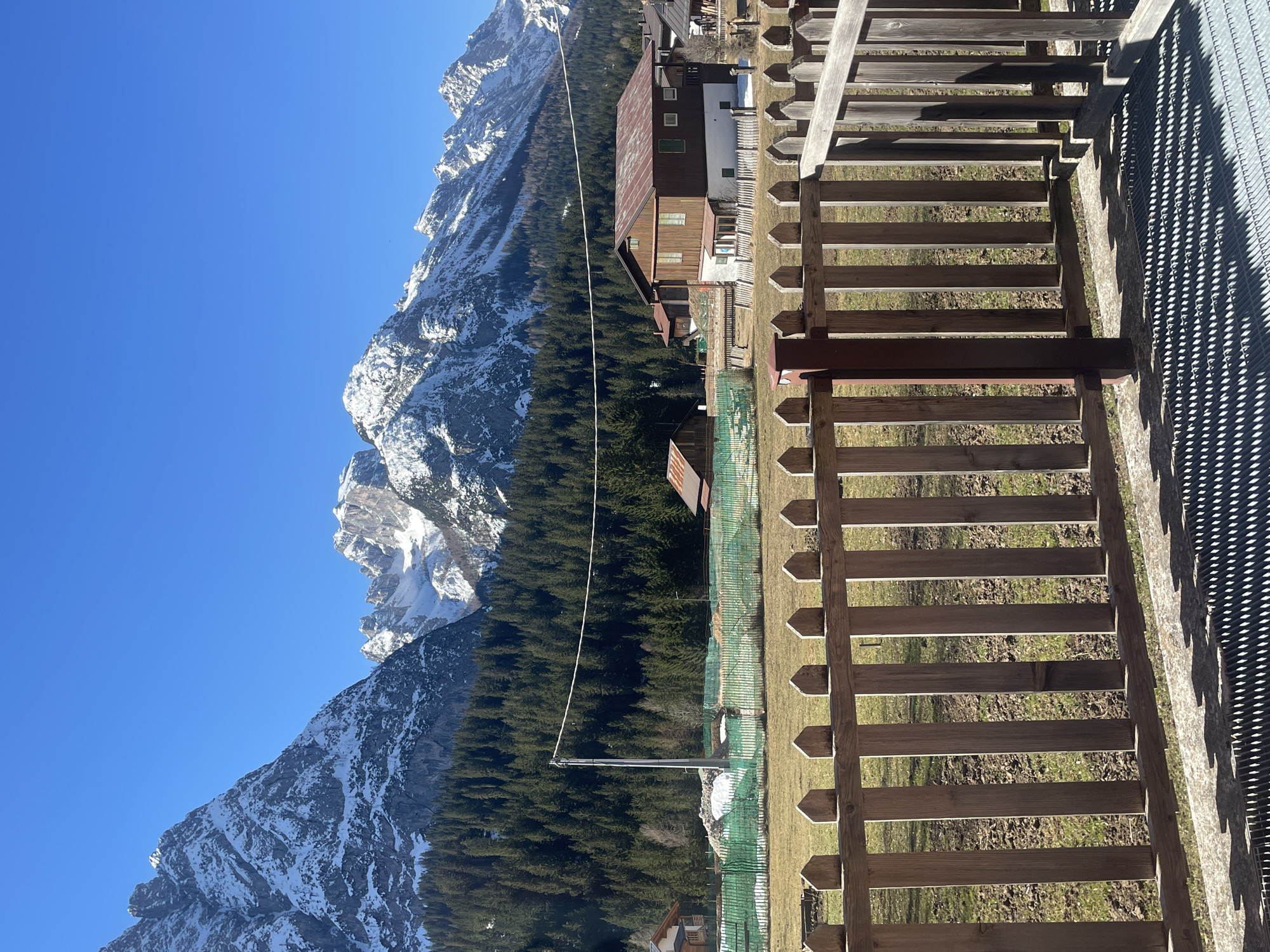 The image shows a landscape view in Sappada, Italy, featuring mountains, forests, and structures. The mountains, partially covered in snow, rise prominently on the left side of the image under a clear blue sky. A dense evergreen forest stretches from the base of the mountains toward the center of the image. Several buildings with rustic architectural styles are nestled within the forest and open areas. A wooden fence runs along the right side of the image, partially obscuring the view. The fence leads to a metal grating. A utility pole with a single wire runs through the center of the image, from the base of the mountains to the buildings. The foreground is dominated by grassy terrain visible through the fence. The location is Sappada / Plodn / Sapade, Italy.