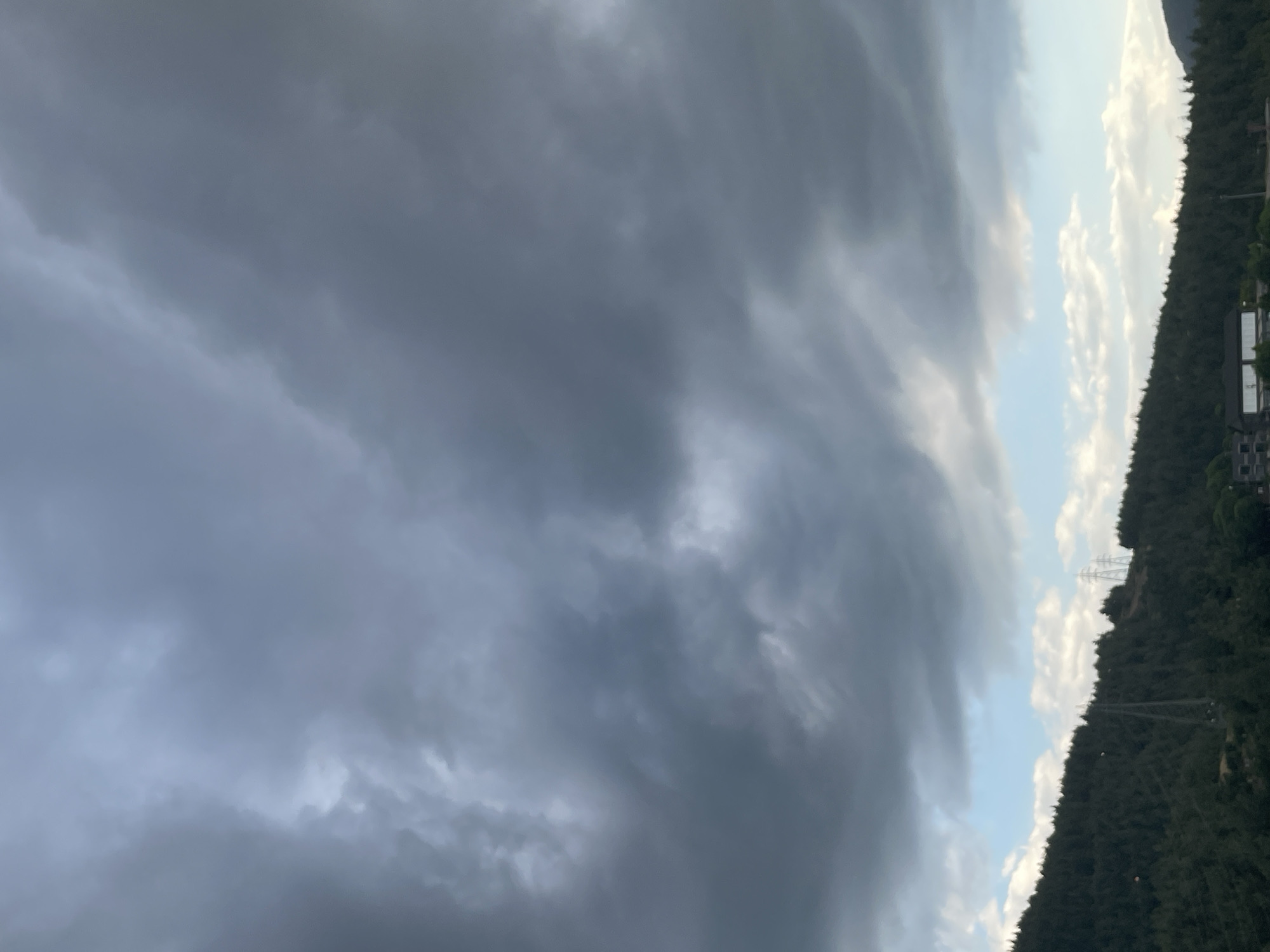 The image shows a dark, cloudy sky over a densely forested hillside in the city of Yunus Emre, Turkey. The clouds are heavy and dark grey, suggesting an impending storm or rain. Below the clouds, there is a strip of lighter sky with some white clouds, and then the dark green expanse of a forested hill. Scattered across the hillside are several tall, slender electricity pylons. At the very bottom of the image, there are some buildings visible, some of which appear to be residential or industrial, nestled amongst the trees. The overall scene is one of a natural landscape under a dramatic sky.