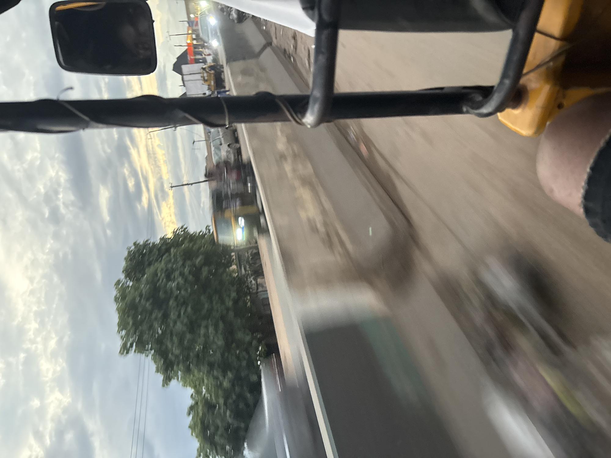 The image captures a street scene in Onitsha, Nigeria, taken from the perspective of someone inside a yellow motorized tricycle, commonly known as a "keke." The foreground is dominated by the tricycle's frame, a black metal bar wrapped with wire, and a portion of its yellow body. A person's leg, clad in black denim shorts with frayed edges, is visible in the lower right quadrant, suggesting the viewpoint of a passenger.
The middle ground of the image depicts a road with a concrete barrier running along its side. The barrier appears weathered and somewhat dirty, with debris visible at its base. Beyond the barrier, traffic is in motion, indicated by motion blur. Several vehicles, including a yellow truck and a few cars, are visible. A distinctive green tree with lush foliage stands out on the left side, partially obscured by the motion blur.
The background reveals a cloudy sky with hints of yellow and orange light, suggesting either sunrise or sunset. A few utility poles with wires are visible against the sky, and buildings with red roofs can be seen in the distance. The overall impression is that of a busy urban environment, possibly during a commute. The scene is characterized by a sense of movement and the typical sights and sounds of a Nigerian city.