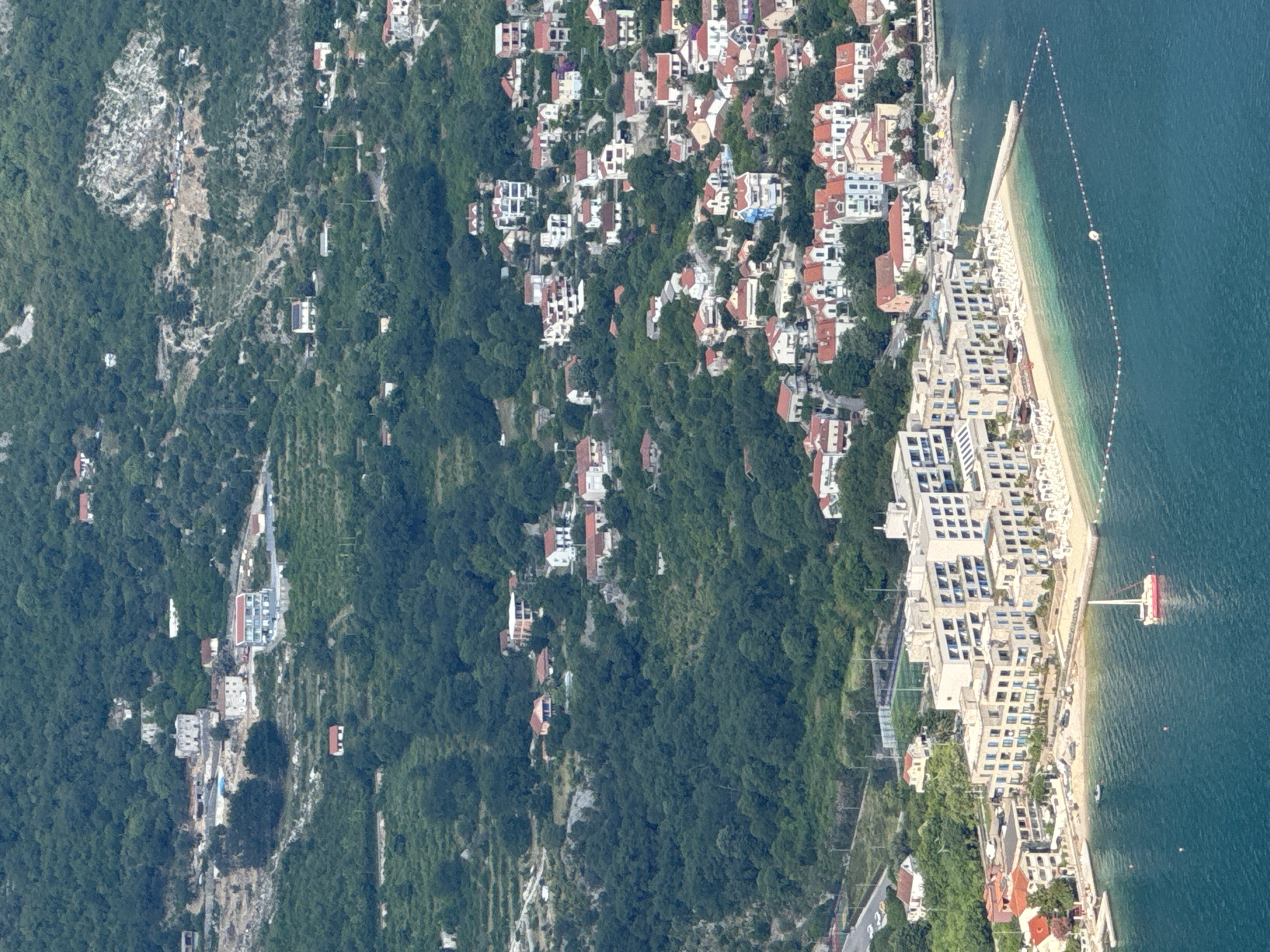 The image shows an aerial view of a coastal town situated on a bay. The town is densely populated with buildings of various sizes and architectural styles, many featuring red-tiled roofs. The buildings are nestled into a steep, green hillside that rises behind the town. In the foreground, a body of turquoise water is visible, with several boats, including a sailboat with a red hull, anchored or moored. A sandy beach is located along the coastline, lined with beach chairs and umbrellas. A modern, multi-story hotel complex with a light-colored facade dominates a section of the waterfront. The overall scene depicts a picturesque coastal settlement with a mix of residential and commercial structures, set against a backdrop of lush vegetation. The location is identified as Strp, Montenegro.