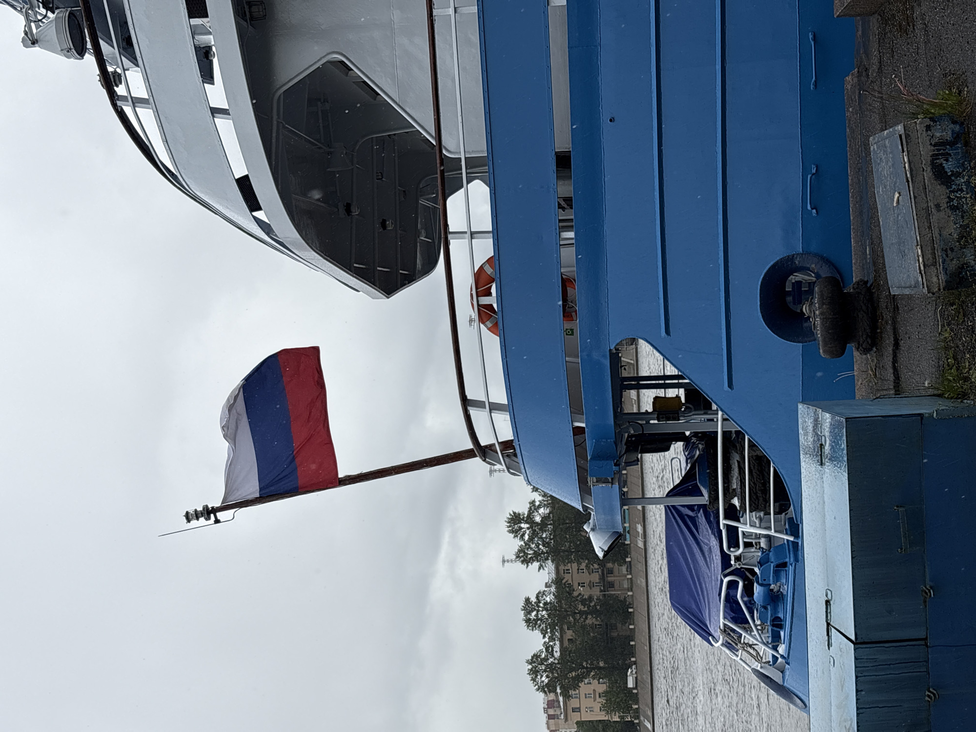The image shows a side view of two boats docked near the embankment of a canal or river in Saint Petersburg, Russia. The boat on the left is grey, while the boat on the right is painted blue. A Russian flag flies from a pole on the left boat. A life preserver is visible on the side of the blue boat. Buildings and trees line the opposite side of the waterway. The sky is overcast.