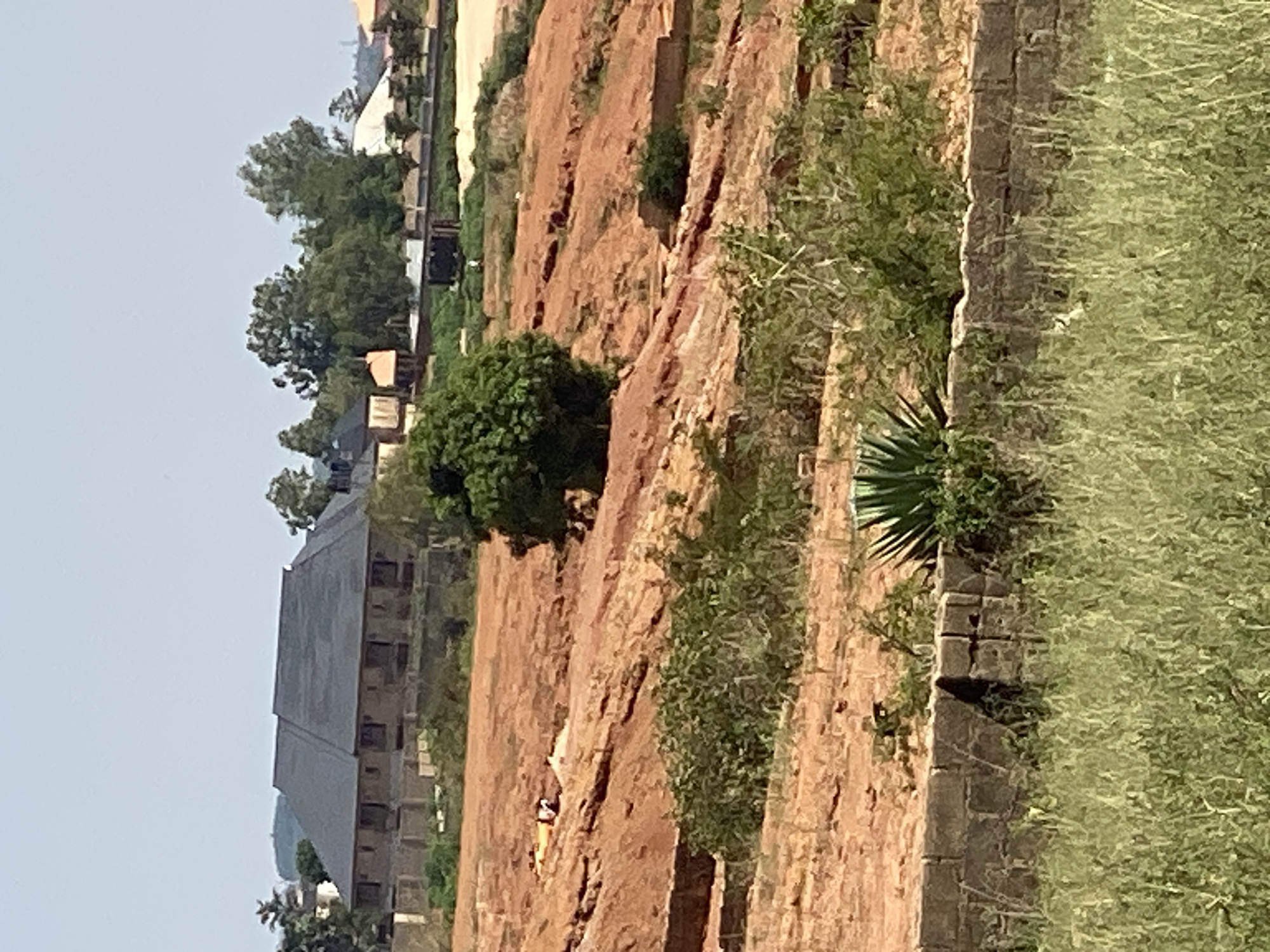 The image captures an outdoor scene in Kwang, Nigeria. In the background, a large, modern house with a dark, sloped roof is visible, constructed with stone or brick. Surrounding the house and extending into the mid-ground are areas of reddish-brown soil, indicative of construction or development. Scattered vegetation, including trees and bushes, dots the landscape. A solitary person, appearing to be laying down on the soil in the mid-ground, is a focal point in the middle of the frame. A low, rough-hewn stone wall runs across the foreground, partially obscuring dense green grass. The sky above is clear and a light blue.