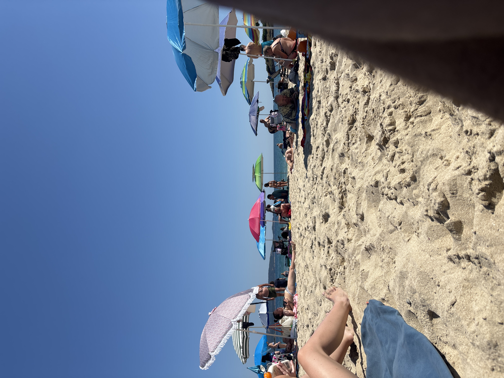 The image depicts a crowded beach on a sunny day. The foreground shows sand with a blue towel and a person's legs and feet. In the midground, several people are sunbathing and relaxing under various colorful beach umbrellas. The umbrellas are a mix of blue, pink, green, and patterned designs. People are scattered across the sand, some lying down, others sitting. In the background, the blue sea meets the clear blue sky, with a distant shoreline visible. The overall scene is one of a typical summer day at the beach in Kiten, Bulgaria.