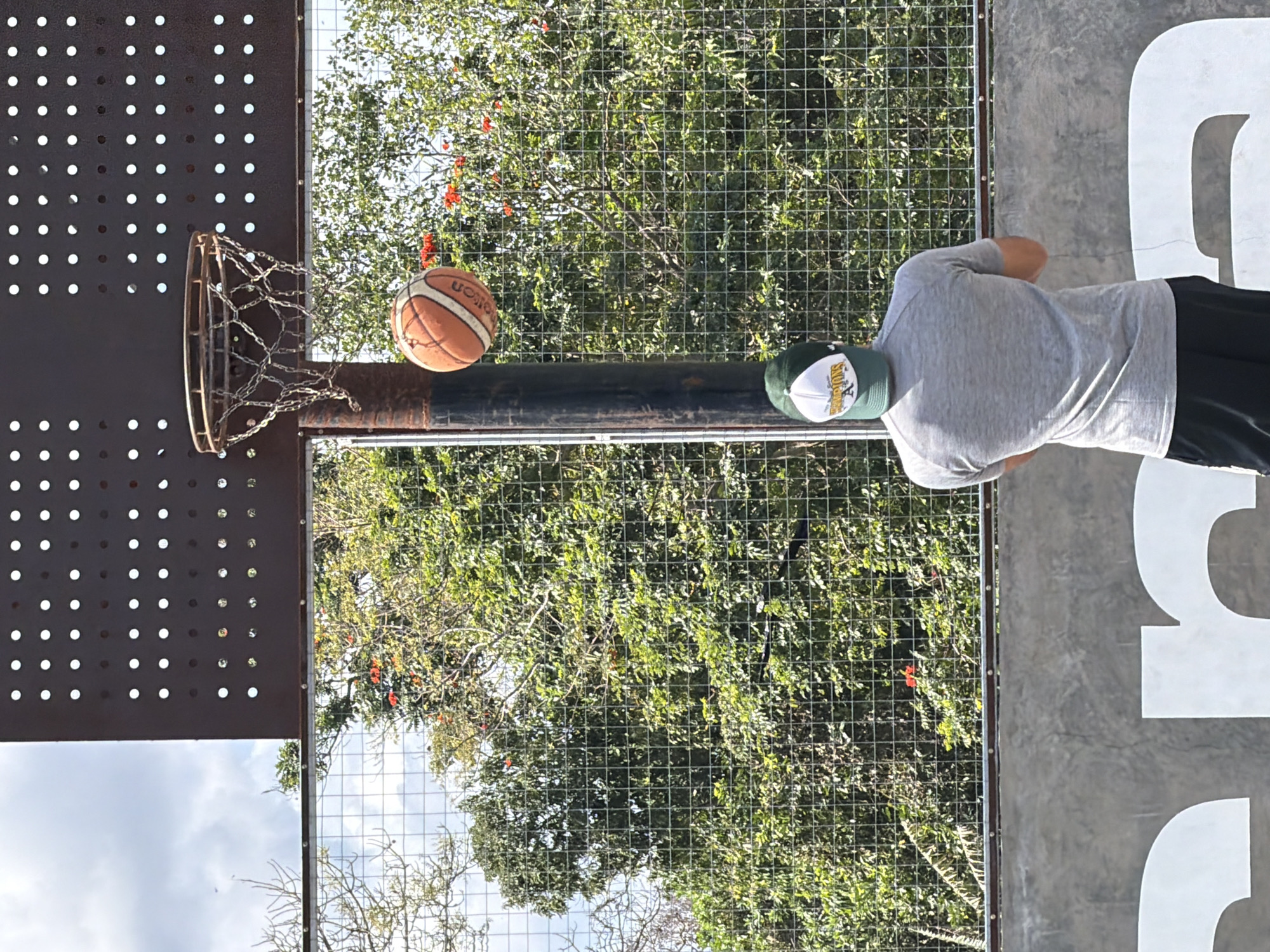 A person wearing a grey t-shirt and black shorts, with a green and white baseball cap on, stands with their back to the camera, facing a basketball hoop and backboard. A basketball is in mid-air, above and to the right of the hoop. The scene appears to be an outdoor basketball court with a chain-link fence in the background, behind which are lush green trees and some red flowers. The backboard is made of a dark, perforated metal. The court has a concrete boundary with large white lettering visible at the bottom of the image. The sky above is partly cloudy. The setting is the outdoor basketball court located in Mengwi, Indonesia.