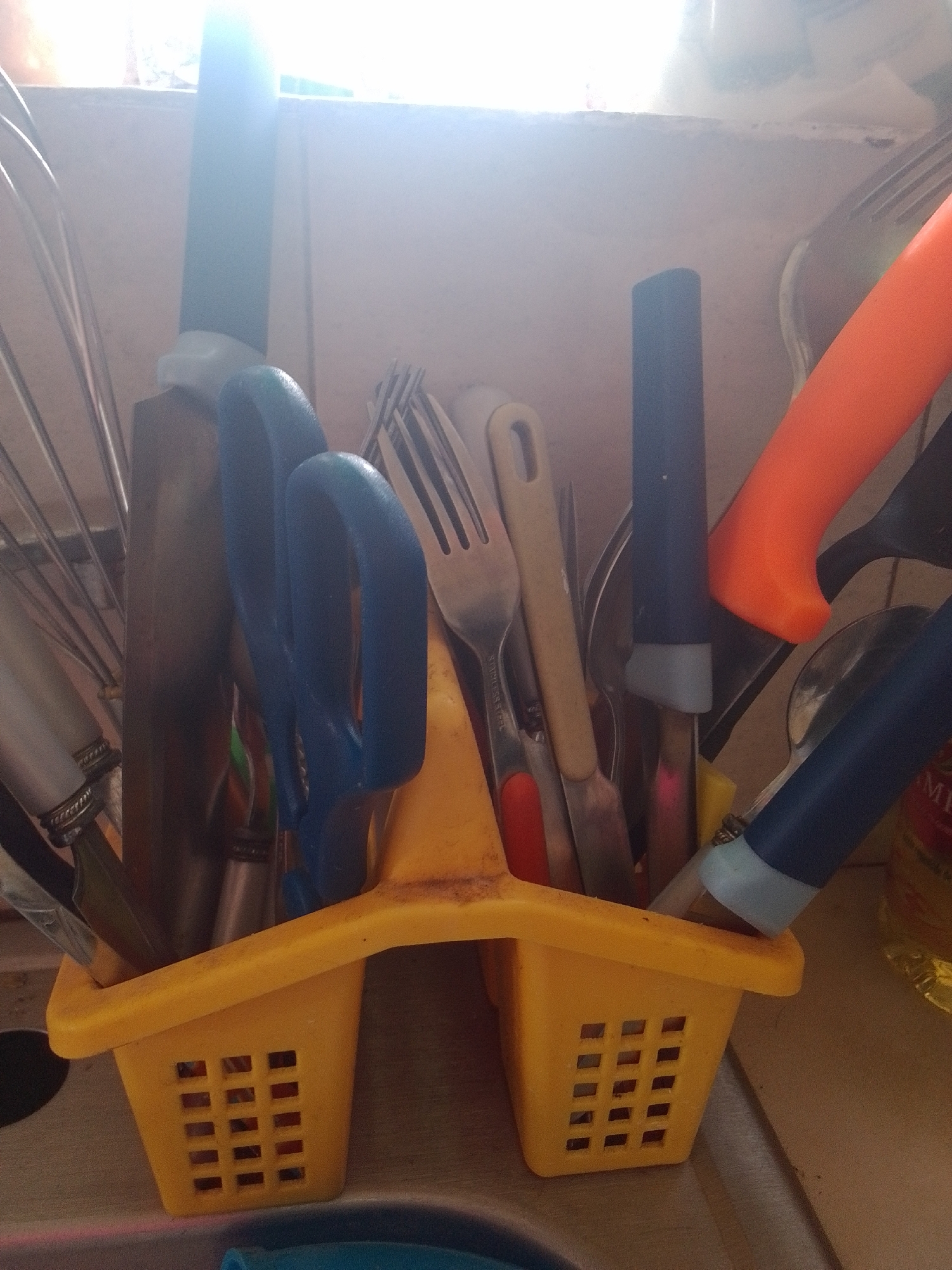 In a kitchen setting, likely in La Tagua, Venezuela, various kitchen utensils are held in a yellow plastic utensil holder. The holder has a rectangular shape with two compartments, each featuring a grid pattern of square openings on the sides.
Among the items visible are a whisk, a large knife with a dark green handle, a pair of blue-handled scissors, and several forks and spoons with metal and light-colored handles. There is also a prominent orange-handled utensil, possibly a knife or spatula, and another utensil with a dark blue handle and a light gray band near its tip. Some of these utensils are partially obscured by others, creating a cluttered but organized appearance.
The lighting suggests it might be daytime, with a bright light source coming from above and behind the utensils, causing some glare and overexposure in the image. The background appears to be a wall or countertop, with a slightly rough texture. The overall impression is of a functional kitchen space where tools are readily accessible. No specific activity or event is depicted, but the arrangement implies recent use or preparation for use. No visible text is discernible in the image.