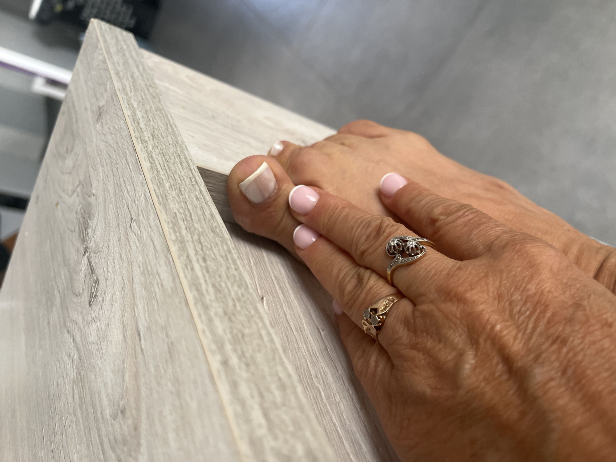 A close-up, top-down shot shows a person's hand interacting with their toes. The hand, adorned with two rings, is positioned over the toes, with the index and middle fingers touching the side of the big toe and the second toe. The toes have a French manicure, with white tips and a pale pink base. The background consists of a light-colored wooden surface, likely a piece of furniture, and a grey floor. The scene appears to be indoors, possibly in a residential setting. There is no indication of the location in Chambœuf, France.