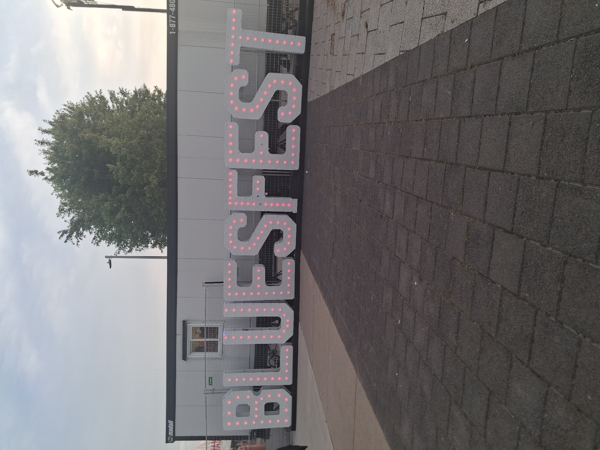 A large outdoor sign with illuminated letters spelling out "BLUESFEST" is prominently displayed. The letters are white with pink lights. Behind the sign is a long, prefabricated building with a small window and a sign that reads "madall". A street lamp is visible above the building, and a large tree is to the right of the building. The foreground consists of a paved walkway made of dark grey interlocking stones, which transitions to a lighter grey stone path on the right. The sky is overcast with some visible clouds. The scene appears to be an outdoor event or venue.