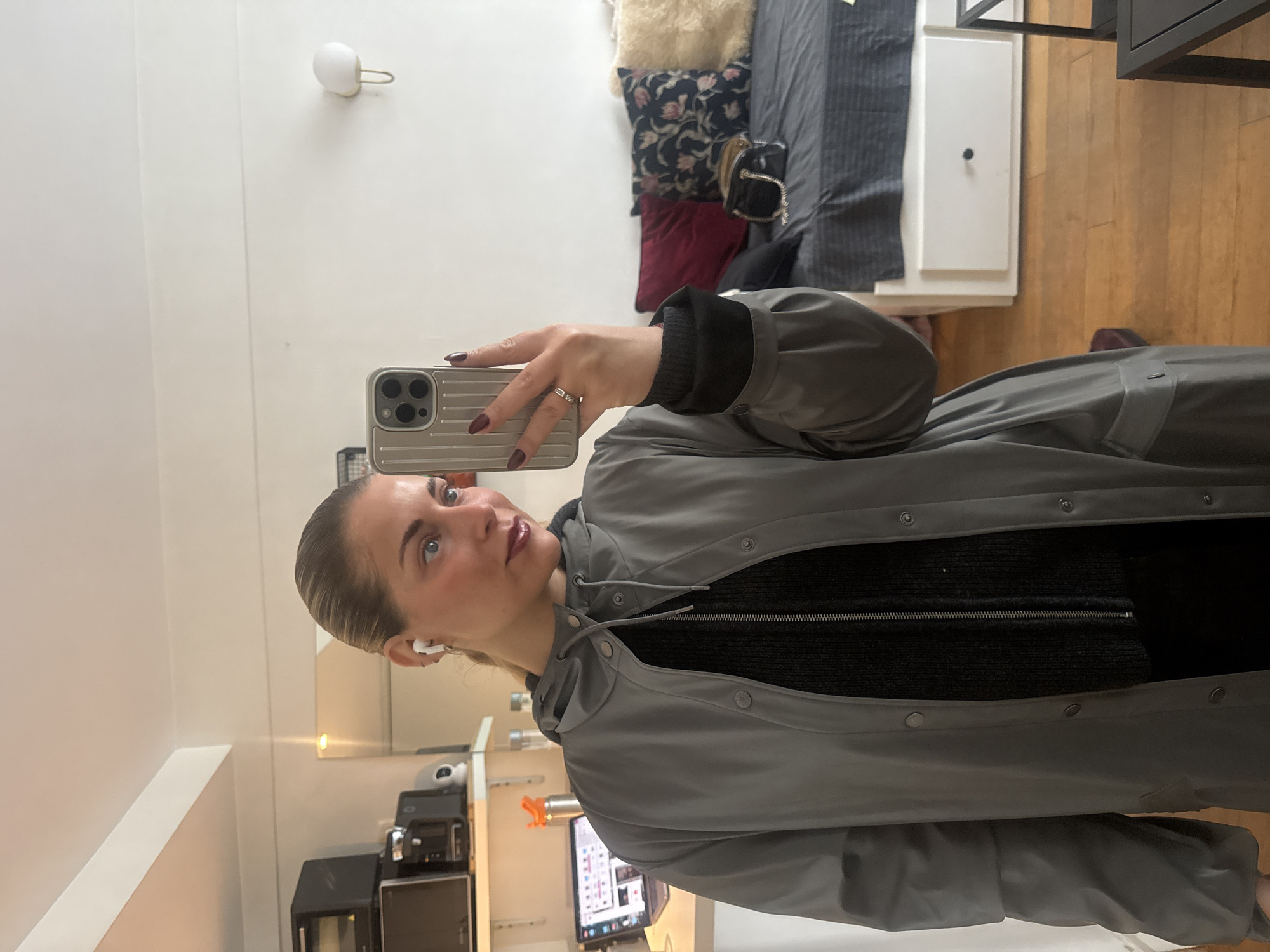A young woman with fair skin and blonde hair is taking a selfie in a well-lit interior. She is wearing a gray jacket over a black top and has an earbud in her left ear. She is holding a silver iPhone with a unique case, showing off her red-painted fingernails and a ring on her finger.
In the background, a desk with a computer monitor displaying content, a microwave, a cabinet, and a small, illuminated wall-mounted light fixture are visible. Above the woman, the corner of a bed with a decorative pillow can be seen, along with a drawer. The presence of a wall-mounted light fixture suggests this is a residential or hotel interior. There is nothing in the frame that would indicate the location is in the city of Paris, France.