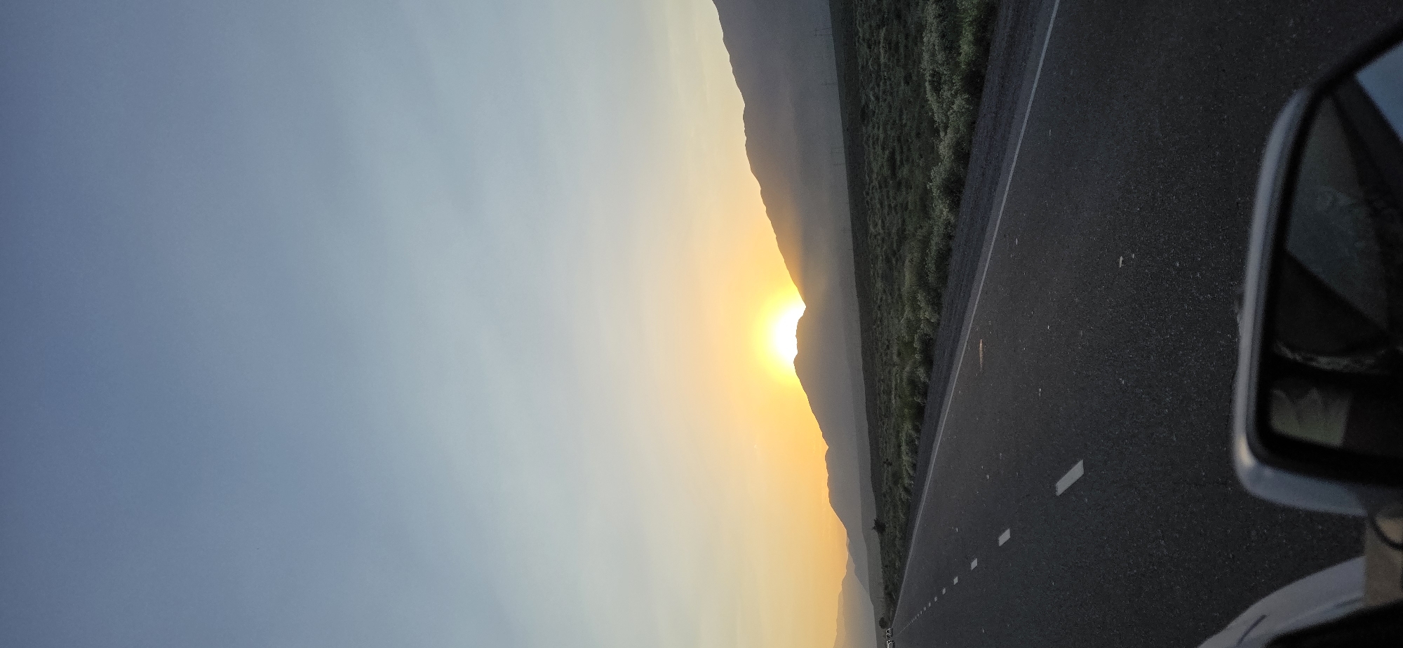 The image captures a scenic view of a highway stretching towards a mountain range at sunrise. The sky is a gradient of pale blue to soft yellow as the sun peeks over the mountains. The road, paved with asphalt, has white dashed lines marking the lanes. To the right of the road, there is a field covered with low-lying green vegetation. In the foreground, a car's side mirror is visible, reflecting the sky and the vehicle's interior. The location is identified as Kok-Moinok - 1, Kyrgyzstan.
