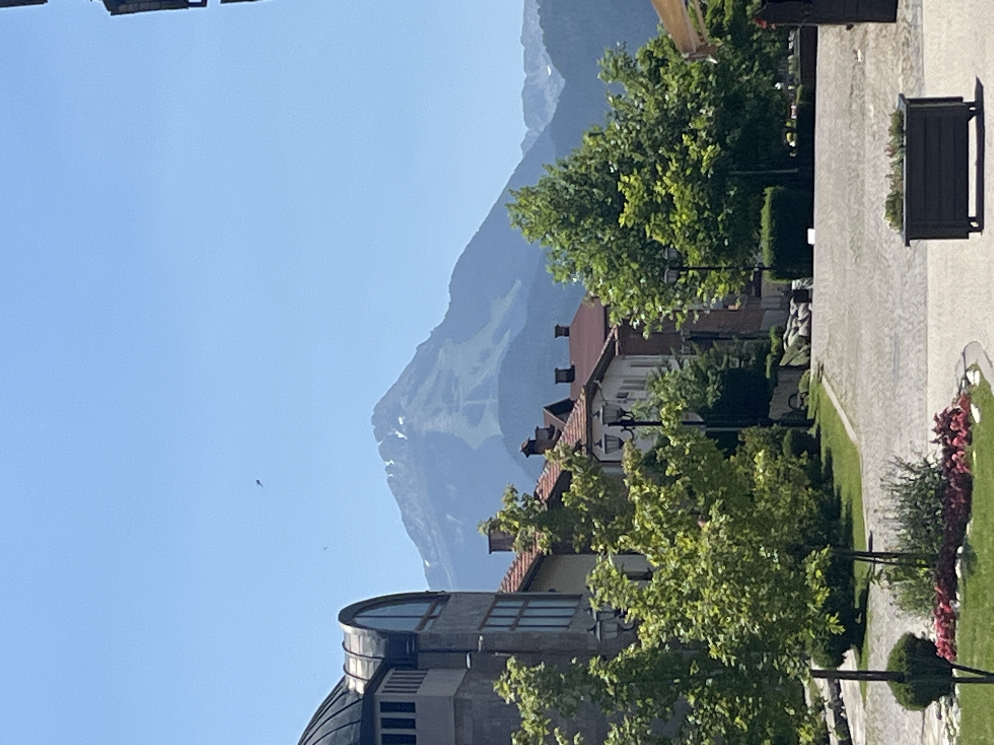 A bright, clear day frames a view of traditional European-style buildings with red-tiled roofs, set against a backdrop of distant, hazy mountains. In the foreground, lush green trees and manicured bushes line a paved walkway. A dark planter box sits on the walkway, filled with small plants. The architecture features a large, grey building with a distinctive rounded, dark roof and arched windows on the left side of the frame. Chimneys are visible on several of the buildings. The sky is a vibrant, cloudless blue. The image appears to be taken in the city of Bansko, Bulgaria, a town known for its mountainous landscape and ski resorts.