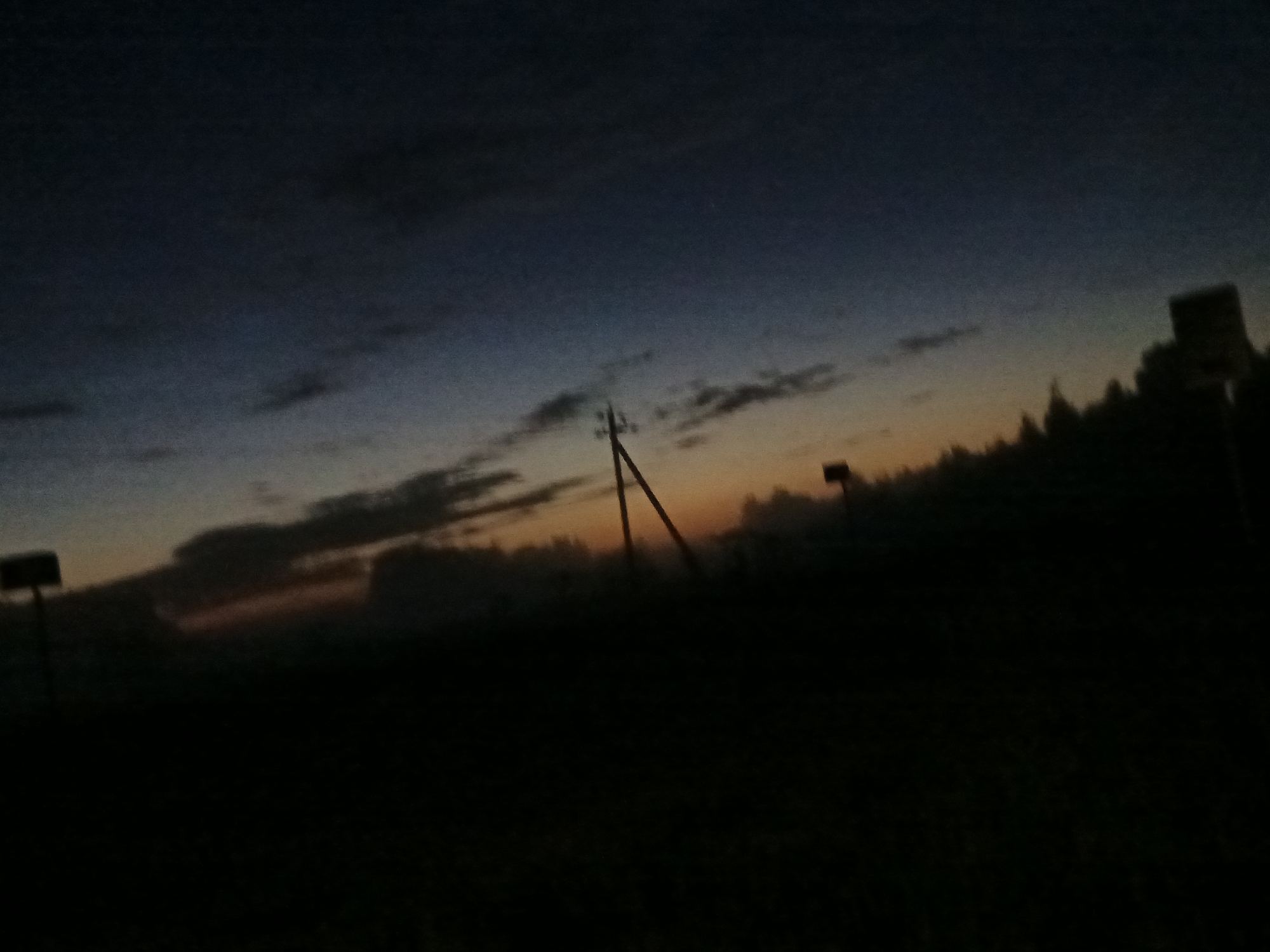The image depicts a twilight sky with a gradient of dark blue fading into orange near the horizon. Silhouetted against the sky are several objects, including a utility pole with wires stretching across the frame, a rectangular sign on a pole to the left, and a darker, more abstract silhouette of trees or a building to the right. The foreground is almost entirely black, suggesting a dark ground cover such as grass or soil, with no discernible details. The lighting indicates it is either dawn or dusk. The scene appears to be an outdoor, possibly rural or semi-rural, location. There are no people or discernible actions taking place. The overall impression is one of quiet solitude.