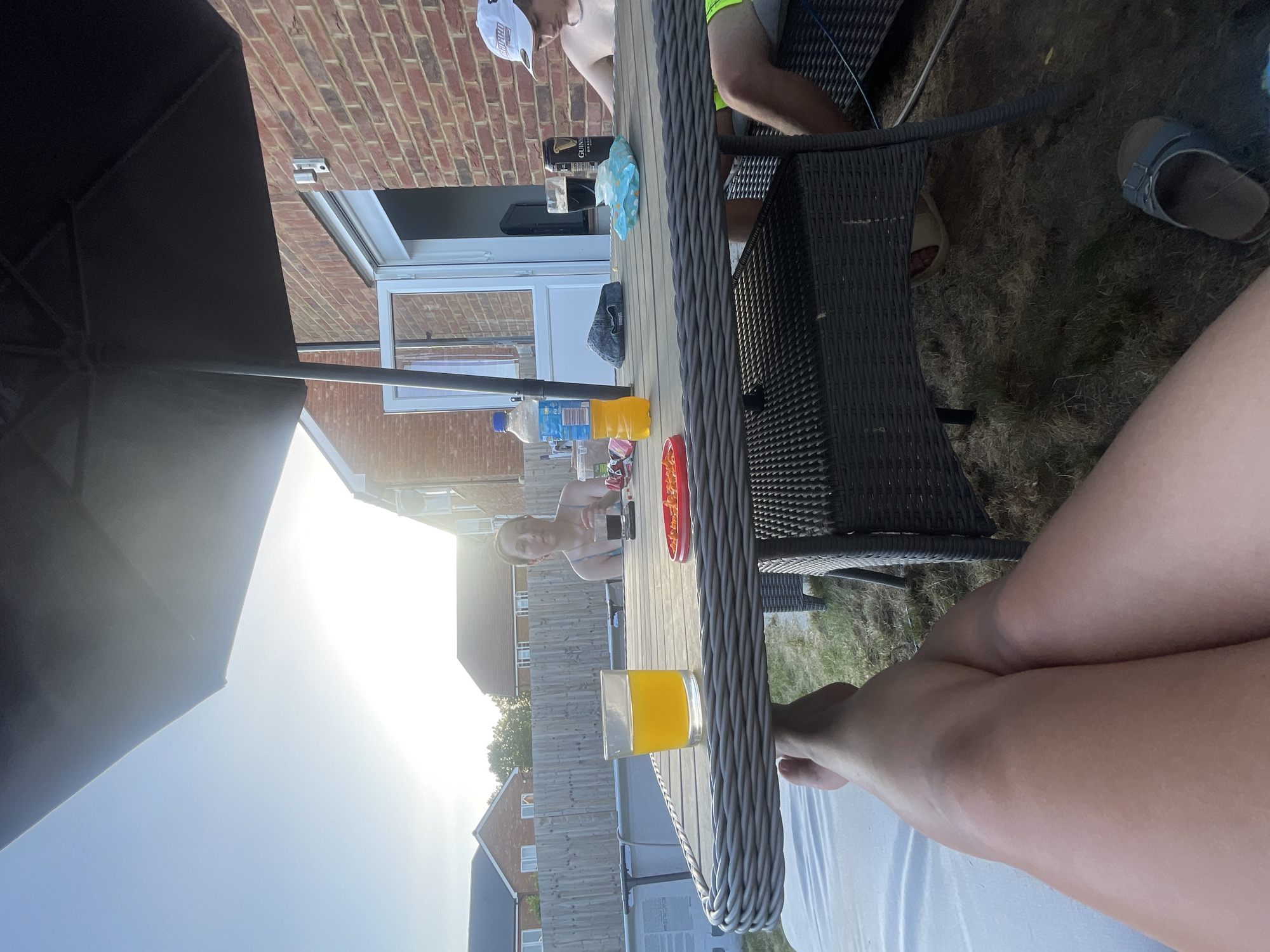 The scene is an outdoor gathering likely occurring in a residential area, possibly in Tonbridge and Malling, United Kingdom. A group of people is sitting around a woven patio table under an umbrella. On the table, there are various drinks, including a glass of orange juice, bottled water, and cans, as well as plates containing food items. The background includes brick buildings, a white-framed door, and residential houses with fences. The foreground features a person's legs resting near the table. The weather appears sunny.
