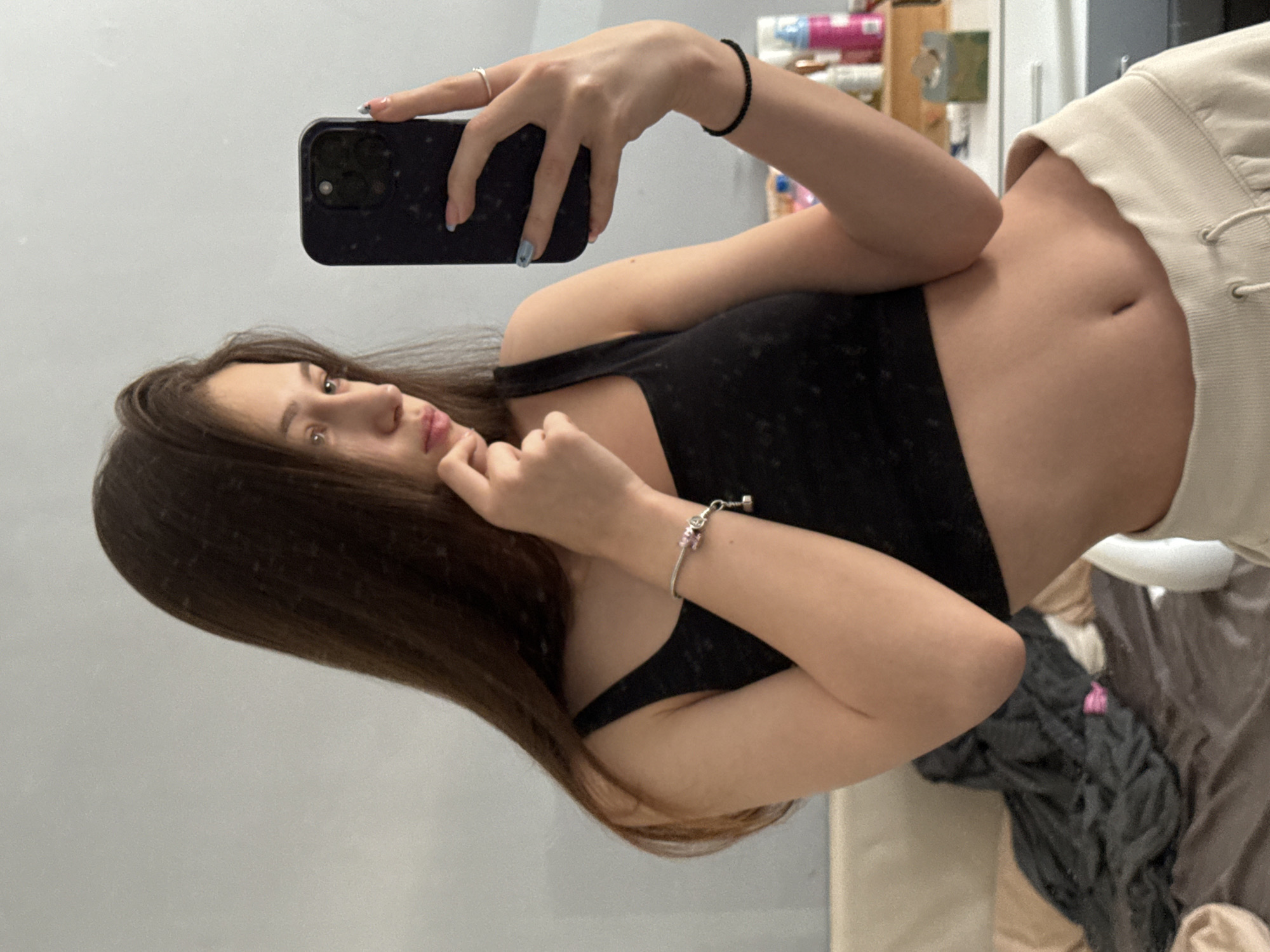 A young woman with long brown hair is posing for a selfie in an indoor setting. She's holding a smartphone in her left hand, and her right hand is near her face in a thoughtful pose. She's wearing a black sleeveless top and light-colored pants or shorts. A silver bracelet is visible on her right wrist, and she has a black elastic bracelet on her left wrist. She is wearing a ring on her left index finger. In the background, there's a cluttered shelf with various personal care items and other objects. The room appears to be a bedroom, based on the presence of bedding in the background. The image does not provide enough information to determine a specific location in Sofia, Bulgaria.