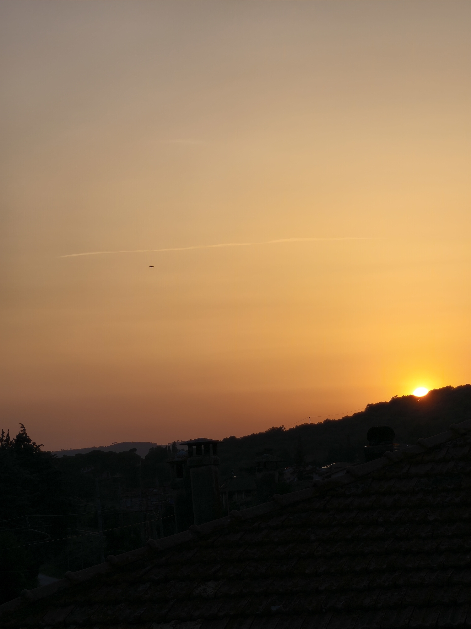 The image captures a sunset in Passignano sul Trasimeno, Italy. The sky is a gradient of warm colors, transitioning from a pale orange at the top to a vibrant yellow near the horizon. A thin contrail from an aircraft is visible in the upper portion of the sky. In the mid-ground, silhouetted against the bright sky, is a hilly landscape with trees. The sun is partially obscured by the crest of the hill, casting a warm glow. In the foreground, the dark silhouette of a tiled roof is prominent, with what appears to be a chimney structure and some trees. The overall scene depicts a peaceful evening view of the town.