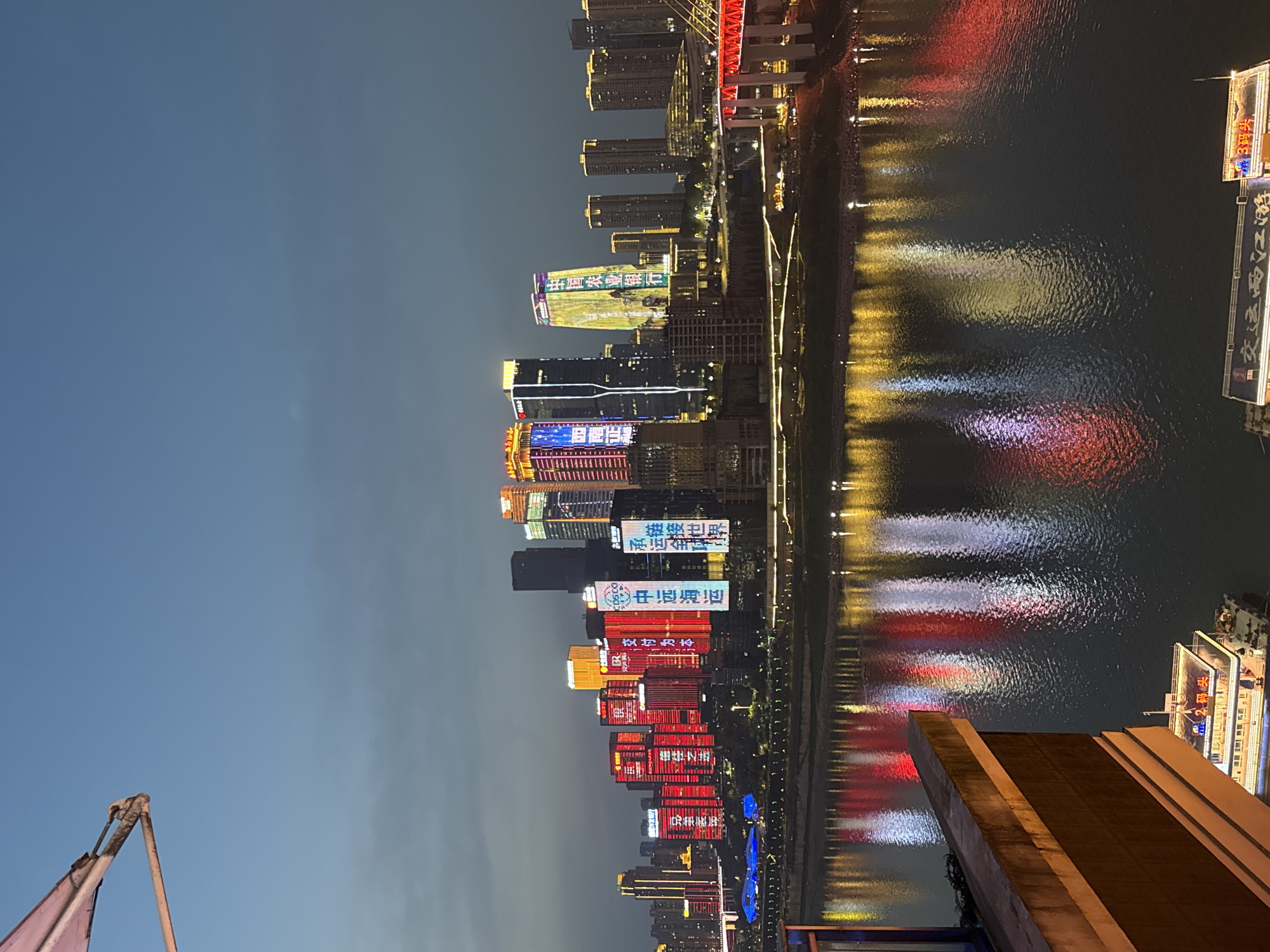 The image shows a nighttime view of the city skyline of Yuzhong District, China, reflected in the river. Tall buildings are illuminated with colorful lights and signage, showcasing Chinese characters. The water reflects these lights, creating streaks of red, blue, yellow, and white. A structure with brown, multi-level roofs is visible in the foreground on the right side of the image. A flag pole and pink flag are visible in the foreground on the left side of the image. Text "2码头" and "3码头" are visible on a building. The sky is dark with a slight gradient.