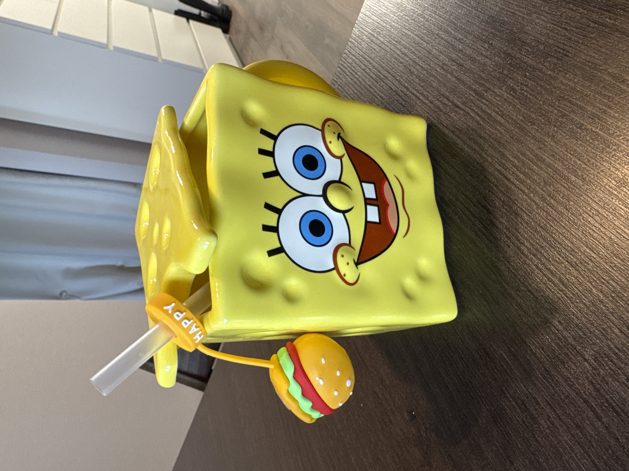 A yellow, square-shaped SpongeBob SquarePants mug with a straw and burger charm sits on a dark brown, wood-grain table. The mug features SpongeBob's face with prominent blue eyes and a wide, grinning mouth. The straw has a yellow "HAPPY" charm attached. A miniature burger charm is also present. In the blurred background, a white cabinet or shelving unit is visible. The image does not provide sufficient information to determine whether it was taken in Chaikovsky, Russia, or any other location.