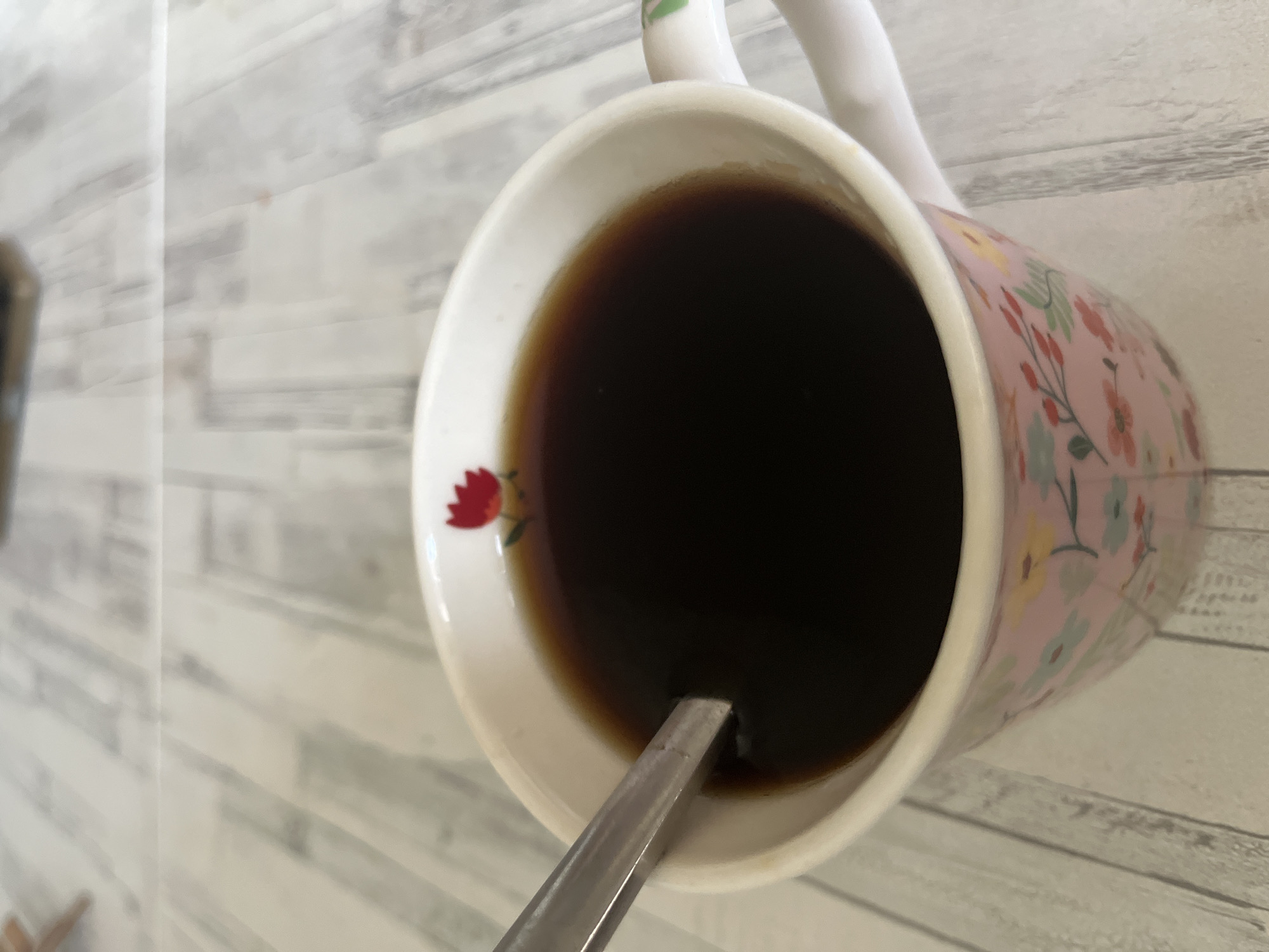 A top-down, close-up shot shows a floral-patterned mug filled with a dark liquid, likely coffee or tea. A spoon is immersed in the liquid. The mug is placed on a surface with a light-colored, wood-grain pattern. No people are visible. The location cannot be determined with the given information beyond the objects and surface in the frame. The city of Chaikovsky, Russia cannot be confirmed as the location.
