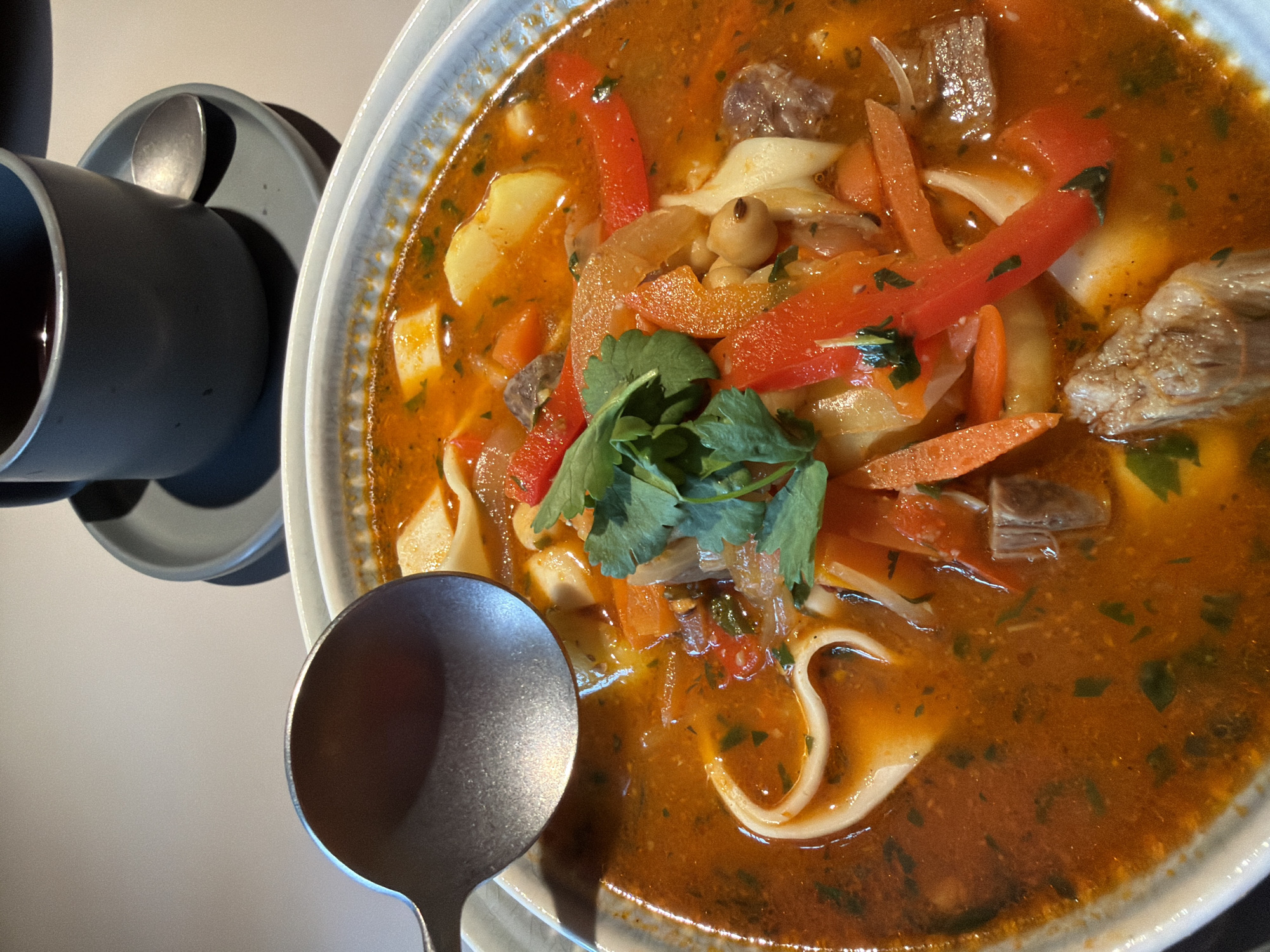 A close-up shot shows a bowl of thick soup containing meat, noodles, carrots, red peppers, and other vegetables, garnished with fresh cilantro. A metal spoon rests on the left side of the bowl. To the left of the bowl is a gray mug and saucer. The overall impression is a meal served in a cafe or restaurant setting. There is no concrete evidence that the image was taken in Chaikovsky, Russia.