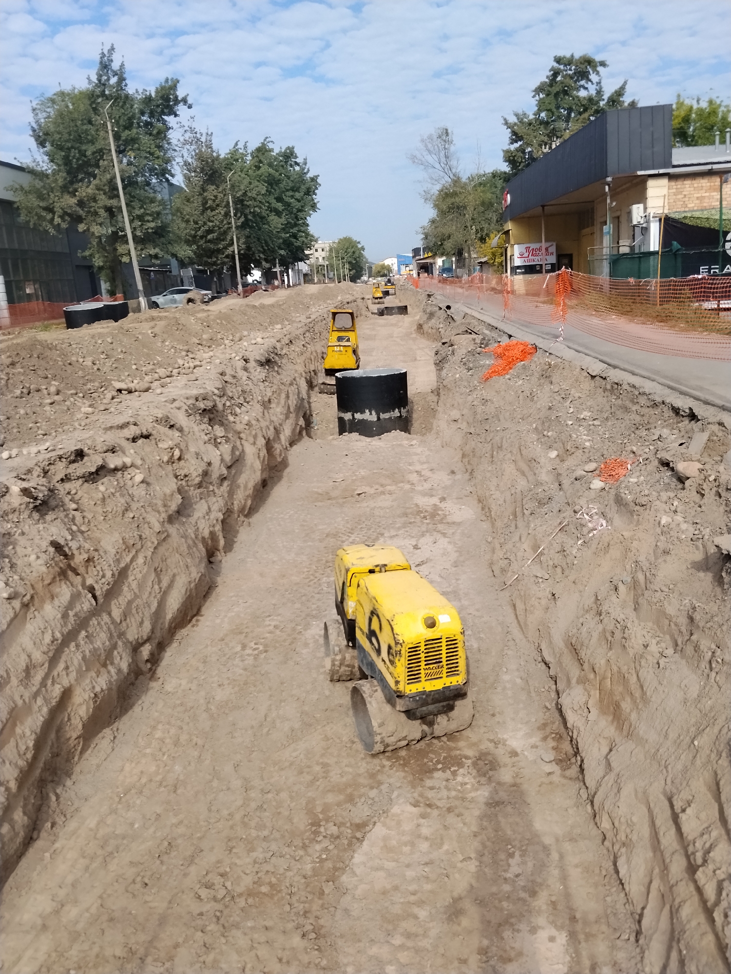 In Bishkek City, Kyrgyzstan, an aerial view captures a construction site with a deep trench running along a paved road. Several yellow construction vehicles, likely compactors, are positioned within the trench. One prominent vehicle in the foreground has the "WACKER" logo visible. Large black cylindrical structures, possibly manholes or pipes, are also present in the trench. The sides of the trench are composed of layered soil and gravel. Orange safety netting and white caution tape delineate the construction area along the road. In the background, trees line the road, and buildings with dark roofs and brick facades are visible. The sky is partly cloudy.