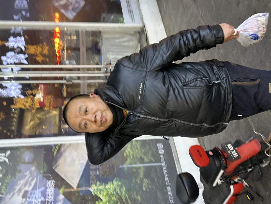 An adult East Asian male is depicted standing on a paved surface, likely a sidewalk, in an urban area of Jiangjin, China. He is facing forward, looking directly at the camera, with a neutral expression. The man has a shaved head and is wearing a black puffer jacket over a black turtleneck or scarf, and dark-colored sweatpants with brown accents. His right arm is bent with the hand positioned behind his head/neck. His left hand holds a transparent plastic bag containing numerous light-colored items, possibly food, and displays a blue logo. To the left and slightly behind the man is the rear section of a red and black electric scooter or bicycle, featuring a red backrest and a fender with Chinese characters "宗田" (Zong Tian) inscribed. The background consists of a glass storefront reflecting bright lights and fragmented images of an advertisement or poster depicting natural scenery and Chinese text. Interior reflections show blurred golden and red/orange lighting fixtures and display items, indicating a commercial establishment. The scene is illuminated, suggesting either evening/nighttime or an artificially lit environment.