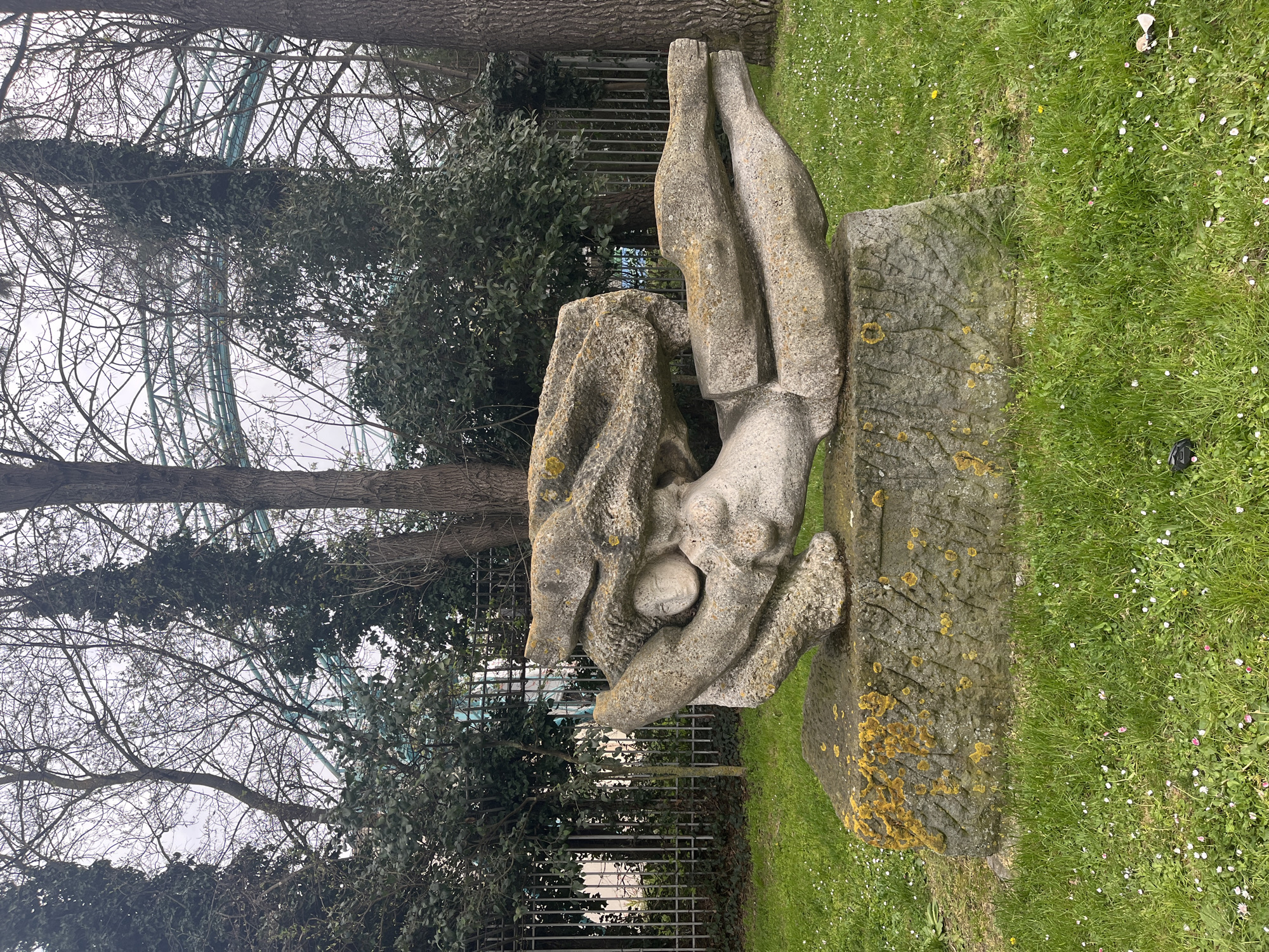 The image shows a stone sculpture of a reclining figure in a park setting. The figure appears to be cradling a round object or part of itself. The sculpture is weathered, with some lichen growth visible. The sculpture rests on a stone base. Behind the sculpture is a green grassy area with white flowers, and beyond that are trees. Through the trees, a metal structure resembling a roller coaster can be seen in the distance. This identifies the location as Burgas, Bulgaria, due to its proximity to the seaside park that features an old coaster. In the background, a fence is also partially visible. The sky is overcast.