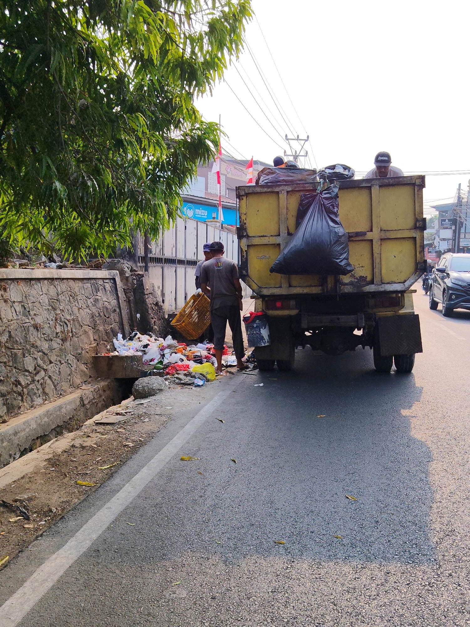 In Purwakarta, Indonesia, a sanitation crew is actively cleaning a roadside drain. Two individuals, dressed in casual clothing, are engaged in the task. One worker, wearing a grey t-shirt and dark pants, is holding a yellow plastic crate filled with debris. He appears to be standing next to a pile of trash that has been removed from the drain. Another worker, wearing a dark shirt and dark pants, is standing nearby, possibly assisting with the cleanup.
Behind them, a yellow garbage truck is parked on the street, its bed filled with collected waste. A large black garbage bag hangs prominently from the back of the truck. The truck appears to be part of the municipal sanitation service.
The scene is set along a street with a concrete drain running alongside the road. The drain is filled with various types of trash, including plastic bags, paper, and other discarded items. A stone wall borders the drain on one side, and trees with green foliage overhang the area. In the background, buildings and power lines are visible, indicating an urban environment. A dark-colored SUV and a motorcycle are also present on the road, suggesting ongoing traffic. The lighting suggests it is daytime, possibly late afternoon due to the warm tone of the sunlight and shadows.
