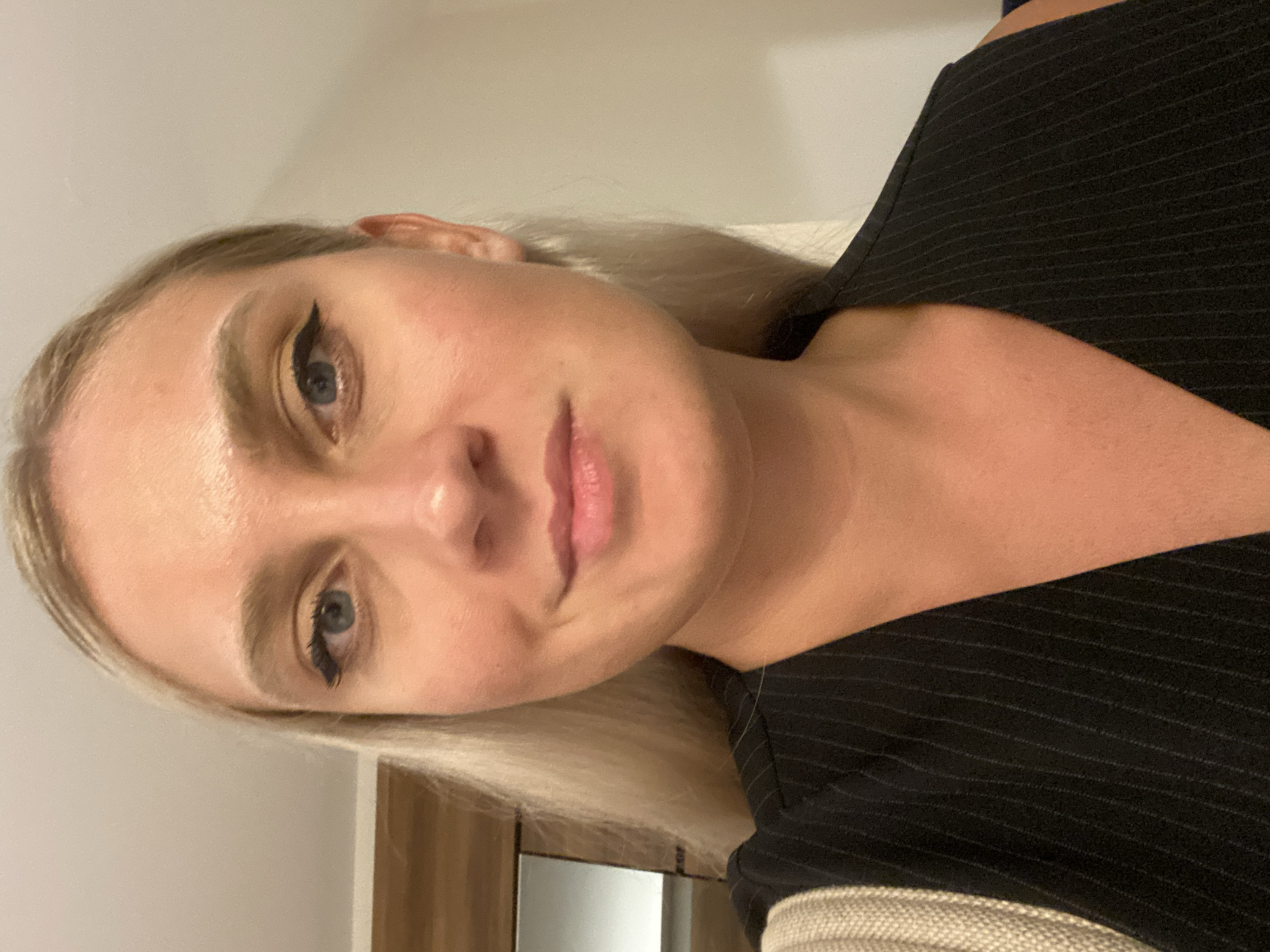 A head and shoulders close-up shot captures a young woman with blonde hair pulled back. She has blue eyes and is wearing makeup, including eyeliner. Her lips are slightly parted, and she has a subtle smile. She is wearing a black pinstripe vest over a tan and white striped garment, possibly a bag strap. The background is a plain, light-colored wall, with a hint of wooden furniture and a framed mirror on the left. The lighting is soft and appears to be indoors. No specific location in Aksu, Turkey can be determined from the visual information provided.