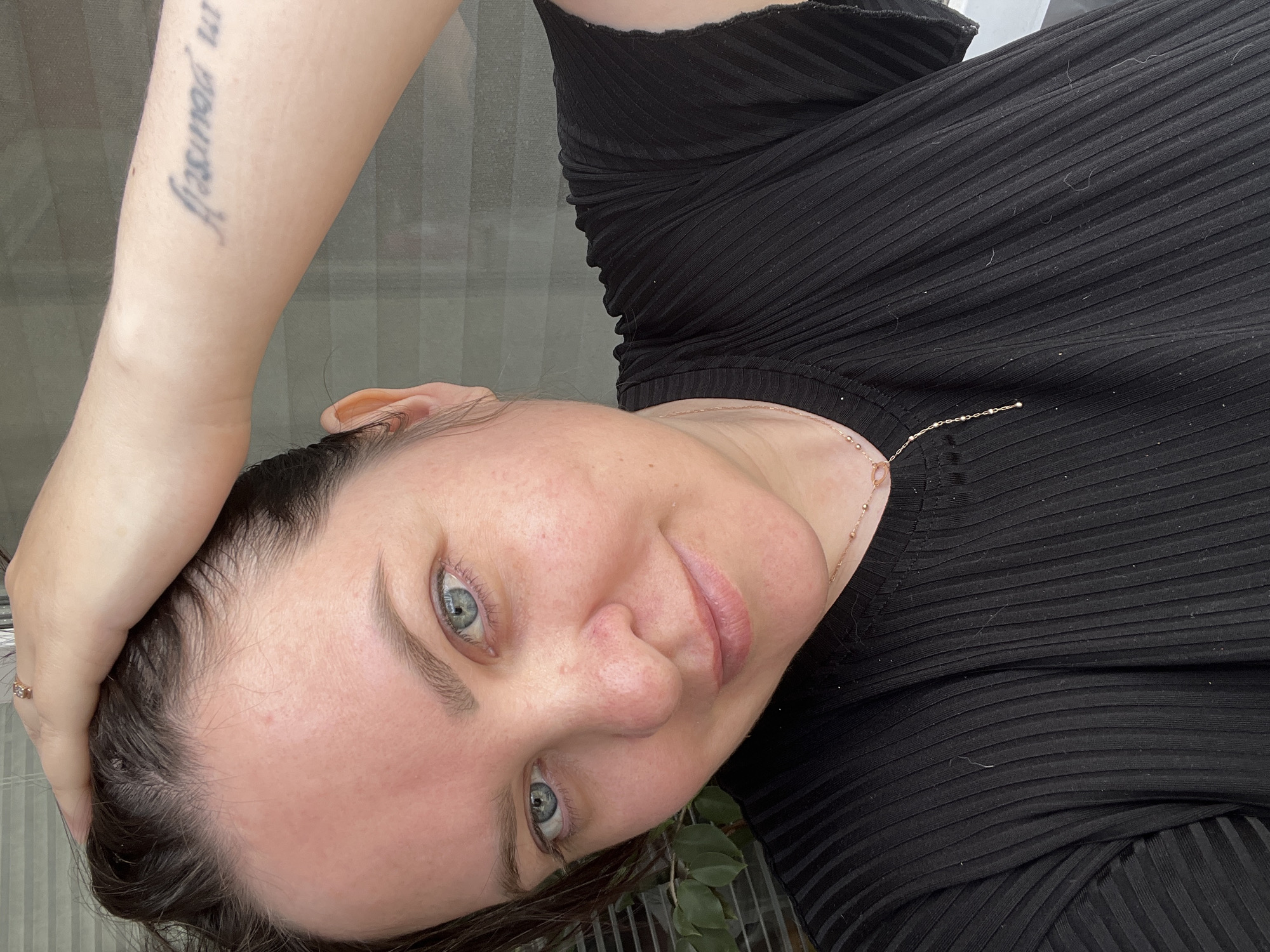 A woman poses with her arm raised to the side of her head, wearing a black ribbed top and a delicate gold necklace. A tattoo is visible on her arm. The backdrop appears to be a window or semi-transparent surface, with greenery partially visible at the bottom of the frame. There is no discernible information to connect the image to Noviy, Russia.
