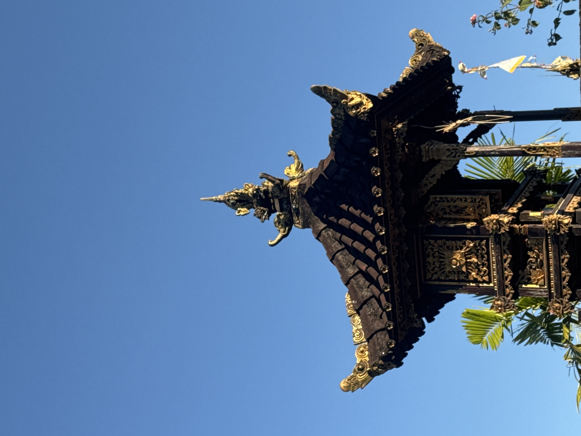 The image shows the detailed, ornate roof of a traditional Balinese building against a clear blue sky. The roof has multiple tiers, covered in dark, weathered tiles. It is elaborately decorated with gold accents, featuring intricate carvings of mythical creatures and patterns along the eaves. A spire-like finial adorns the highest point, also embellished with gold. The building itself appears to be made of dark wood with carved panels, visible beneath the roofline. Palm fronds are visible to the left, and a small flag and flowering plant are seen to the right. The setting suggests a tropical location. This structure is likely a temple or part of a larger complex, potentially in Kuta Selatan, Indonesia, given the architectural style. There are no people or discernable actions in the image.