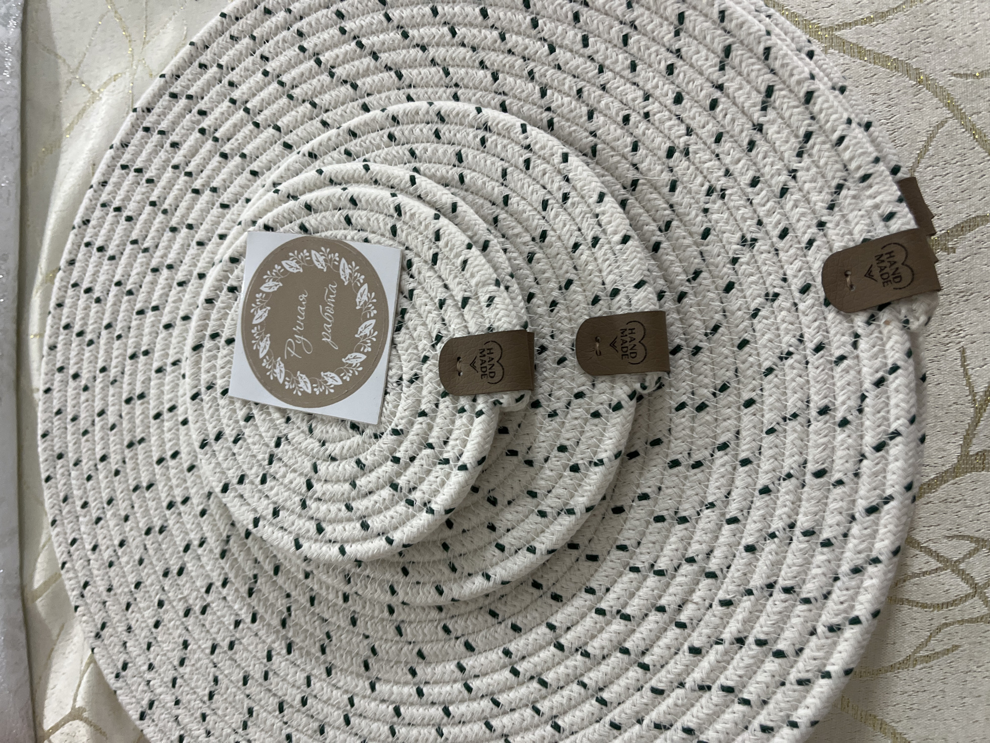 The image shows three white woven circular placemats stacked on top of each other. The placemats have a repeating pattern of small dark green dashes along the woven strands. A circular sticker with a floral border and text in a script font is placed on the top placemat. Below the sticker, three small brown leather tags, each imprinted with "HAND MADE" in a circular shape, are attached to the edges of the placemats. The background appears to be a textured fabric surface with a subtle golden pattern. There are no people visible in the image. The scene is indoors, with soft lighting. The location in Hrodna, Belarus is not discernible from the image.