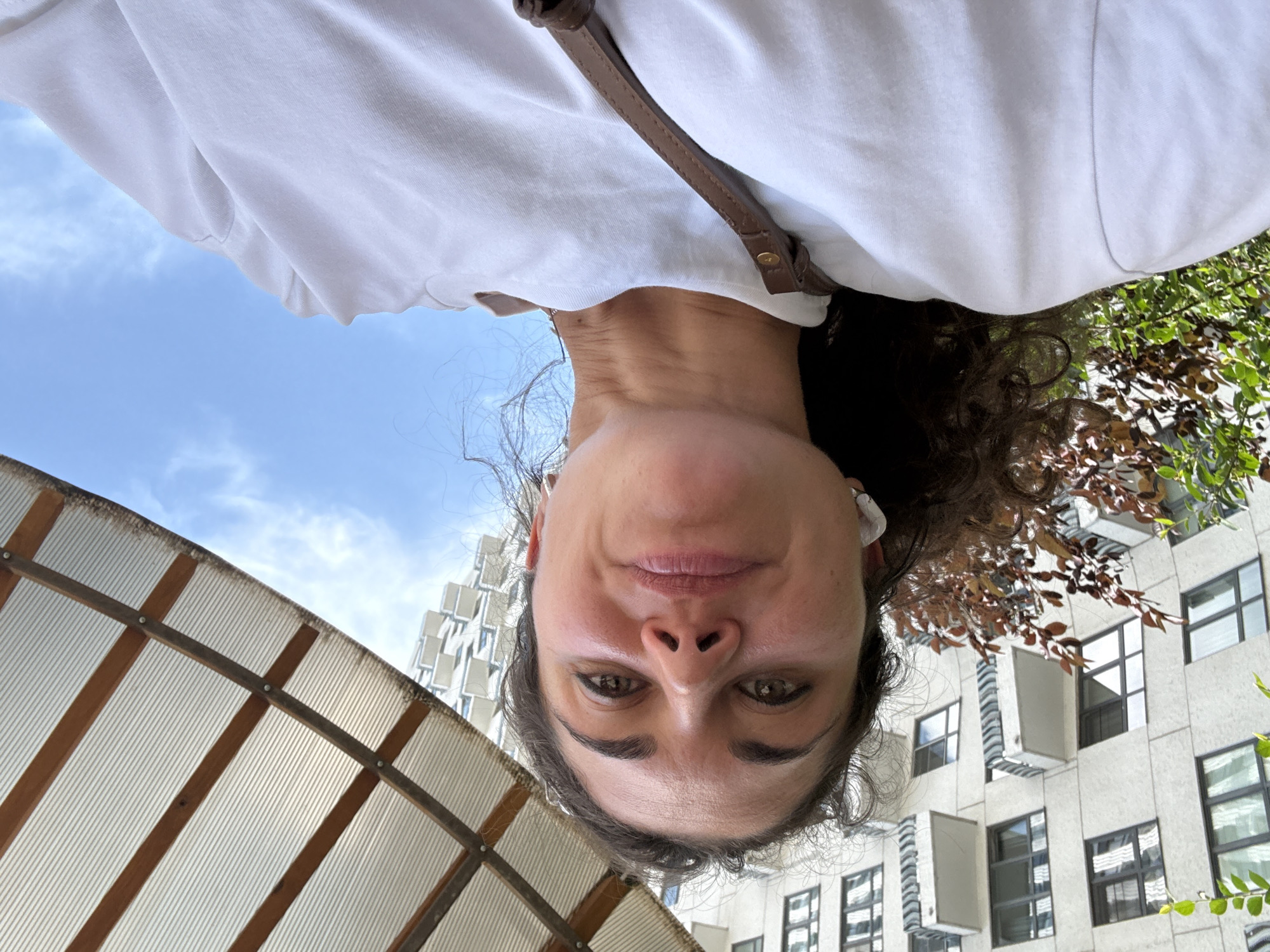 A woman is taking a selfie outdoors with a building and sky in the background. She is wearing a white t-shirt and has dark curly hair. There is a brown strap visible over her shoulder. The building is modern with many windows and balconies. The sky is blue with some clouds. The image appears to be taken at an upward angle, suggesting she is looking up at the camera.