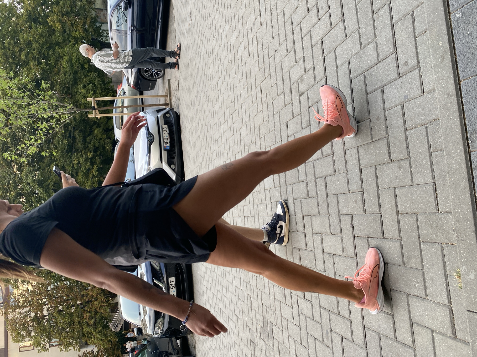 An outdoor, eye-level, full shot captures two people walking side-by-side on a sidewalk paved with grey rectangular stones, set in a herringbone pattern. To the left, a woman with her back to the camera is wearing a black t-shirt and black athletic shorts. She is wearing a blue beaded bracelet on her left wrist and pink athletic shoes. Her right leg is extended forward, and her left leg is slightly bent. Her hair is pulled back into a ponytail.
To the right, a man with grey hair and glasses is wearing a short-sleeved, patterned shirt and dark pants. He is wearing dark sandals and holding a smartphone in his right hand. He is looking down at his phone.
In the background, several parked cars are visible. On the left, a silver sedan with license plate "NBU 138" is partially visible. A black sedan is parked to its right. Further in the background, a man in a light-colored shirt and dark pants, possibly the same man as the one on the right, is walking away from the camera. Trees with green foliage are visible behind the cars. The scene appears to be in a city, possibly Chișinău, Moldova, given the license plate format, although this cannot be definitively confirmed from the image alone.