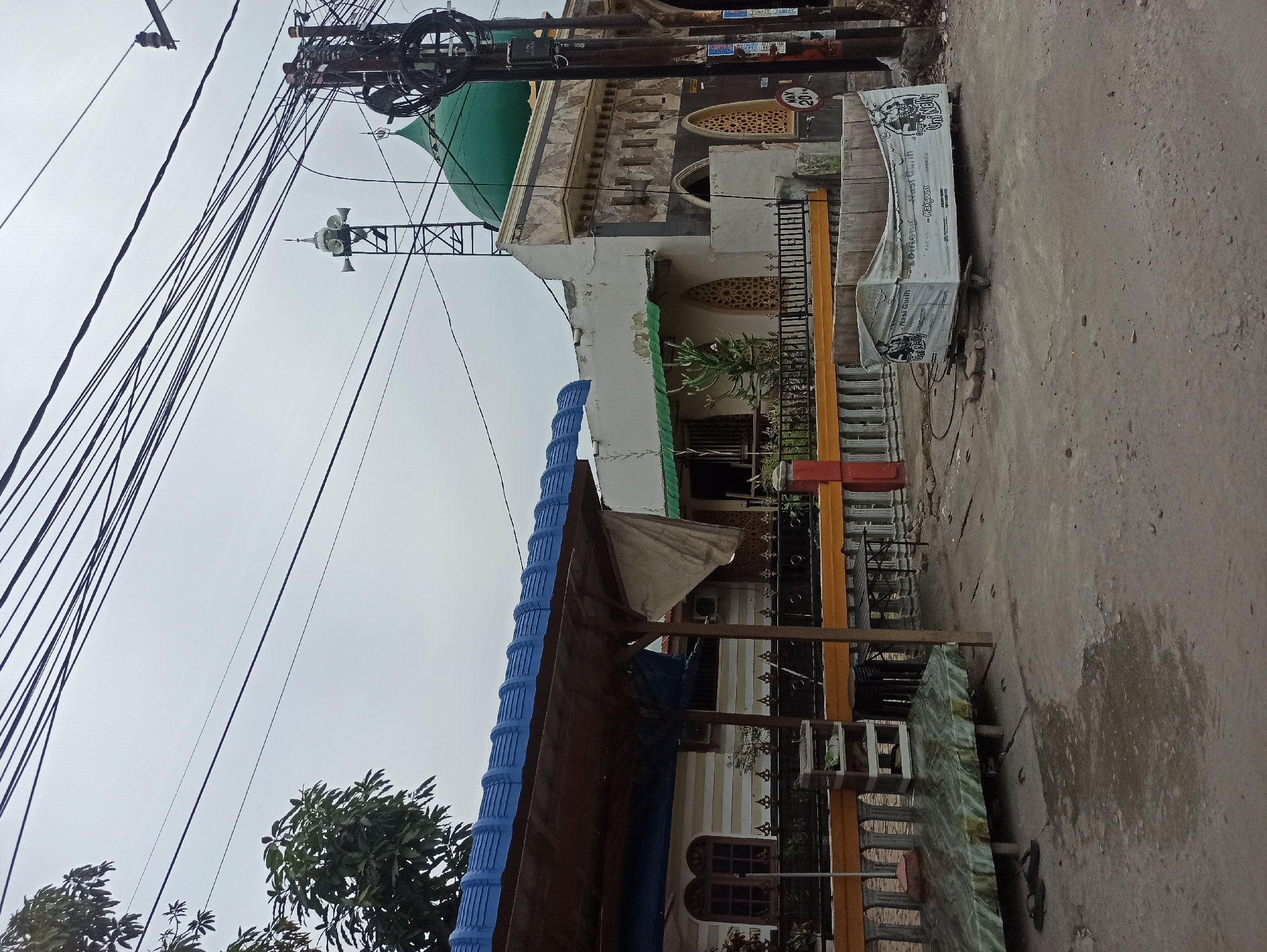 In the city of Medan, Indonesia, an overcast sky looms above a street scene. To the left, a blue-roofed structure with a brown awning covers a seating area with a green and white patterned mat and some flip-flops. A fence, adorned with a yellow and white railing, separates this area from a building with striped walls and two arched windows. Further to the right, a mosque with a green dome and intricate architectural details stands prominently. A tall tower with speakers is situated near the mosque. The street itself is paved with concrete, showing signs of wear and tear, with some debris scattered around. A white cart, draped with a tarp featuring a cartoonish illustration and Indonesian text, sits in the foreground, suggesting a food stall or temporary vendor. Electrical wires crisscross the sky, indicating the urban setting. A "MAX 20 km/h" sign is visible on a pole near the mosque.